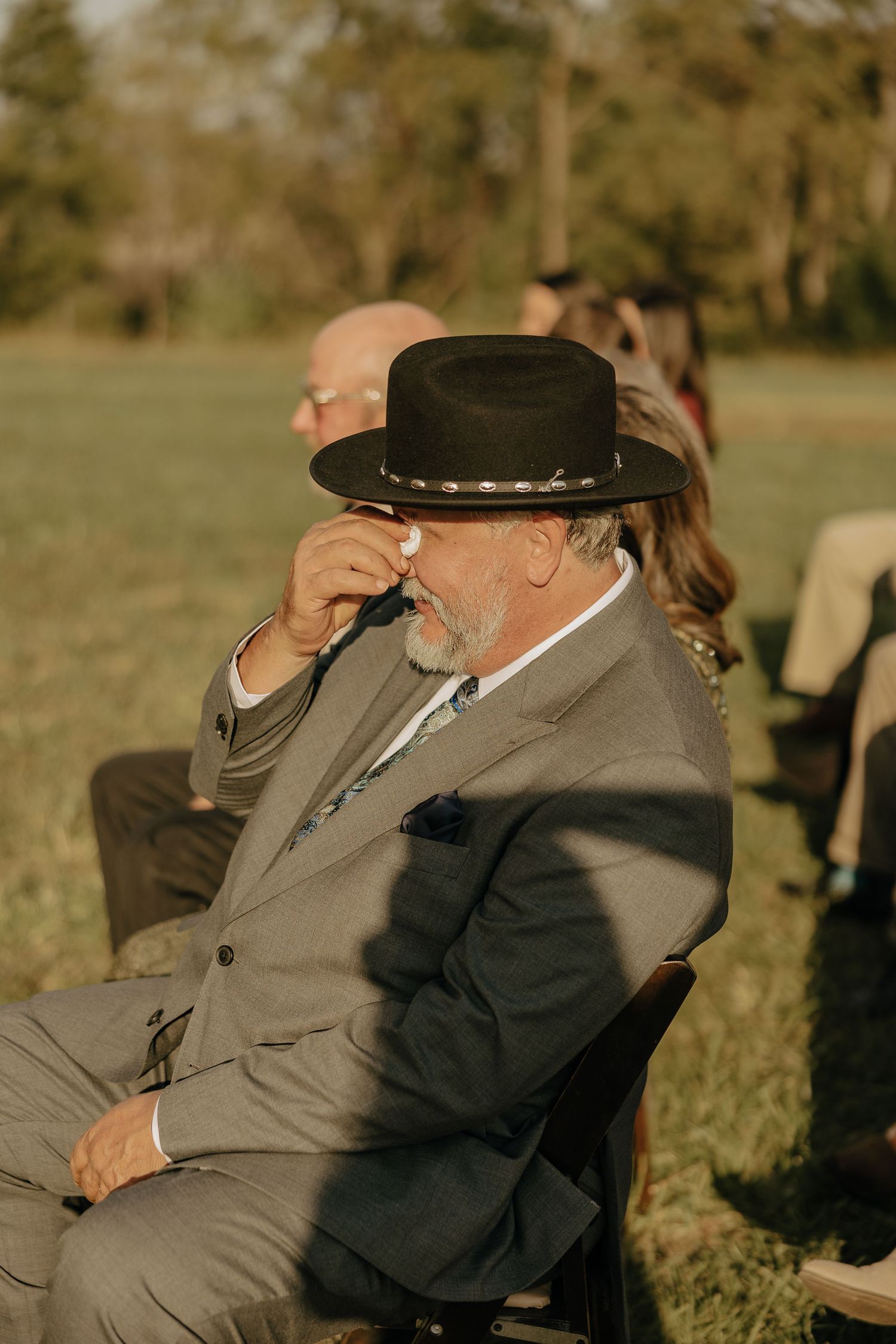 A person in a suit and cowboy hat sits outdoors during a golden hour wedding celebration.