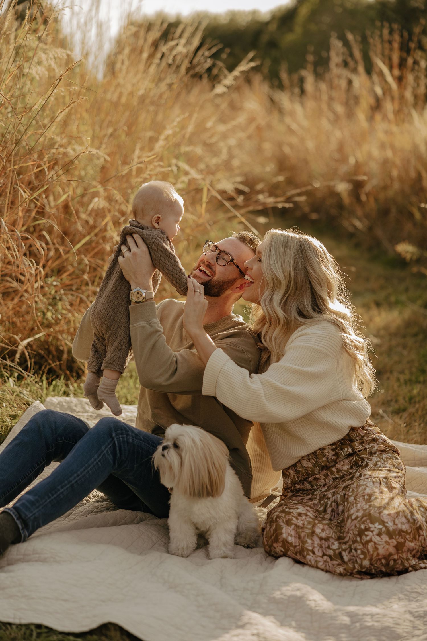 Candid family moments captured on a white blanket with their small white dog in golden sunlight.