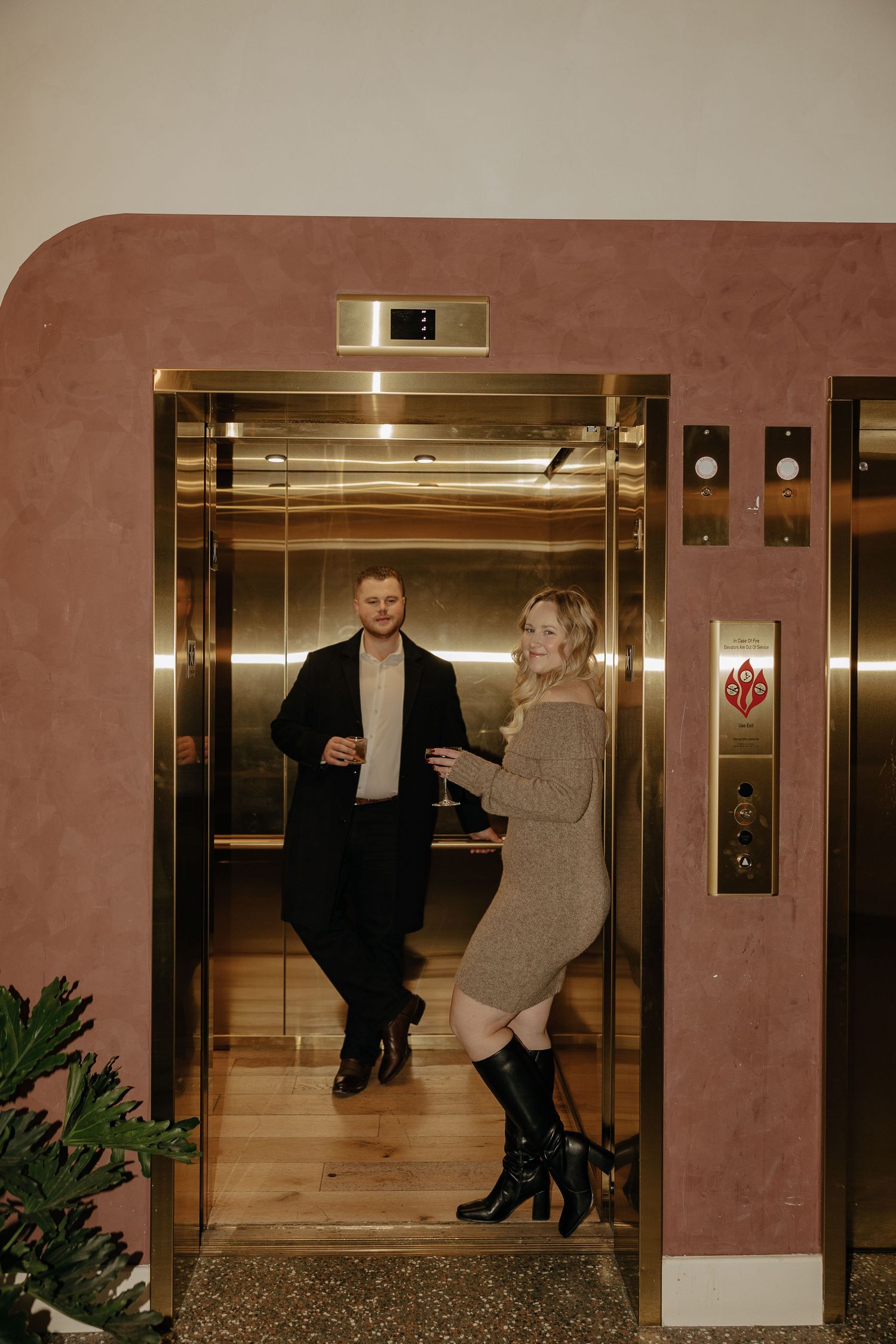 A couple playfully poses in an elevator lobby with warm lighting and brown walls.