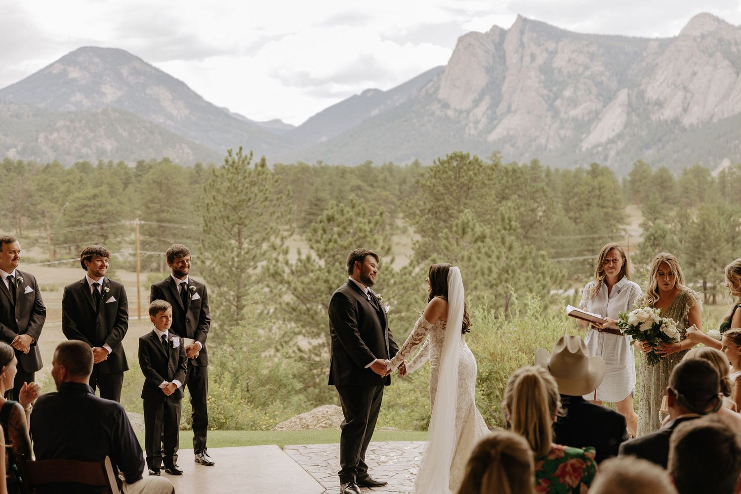 An outdoor wedding ceremony takes place with mountain peaks as a dramatic backdrop.