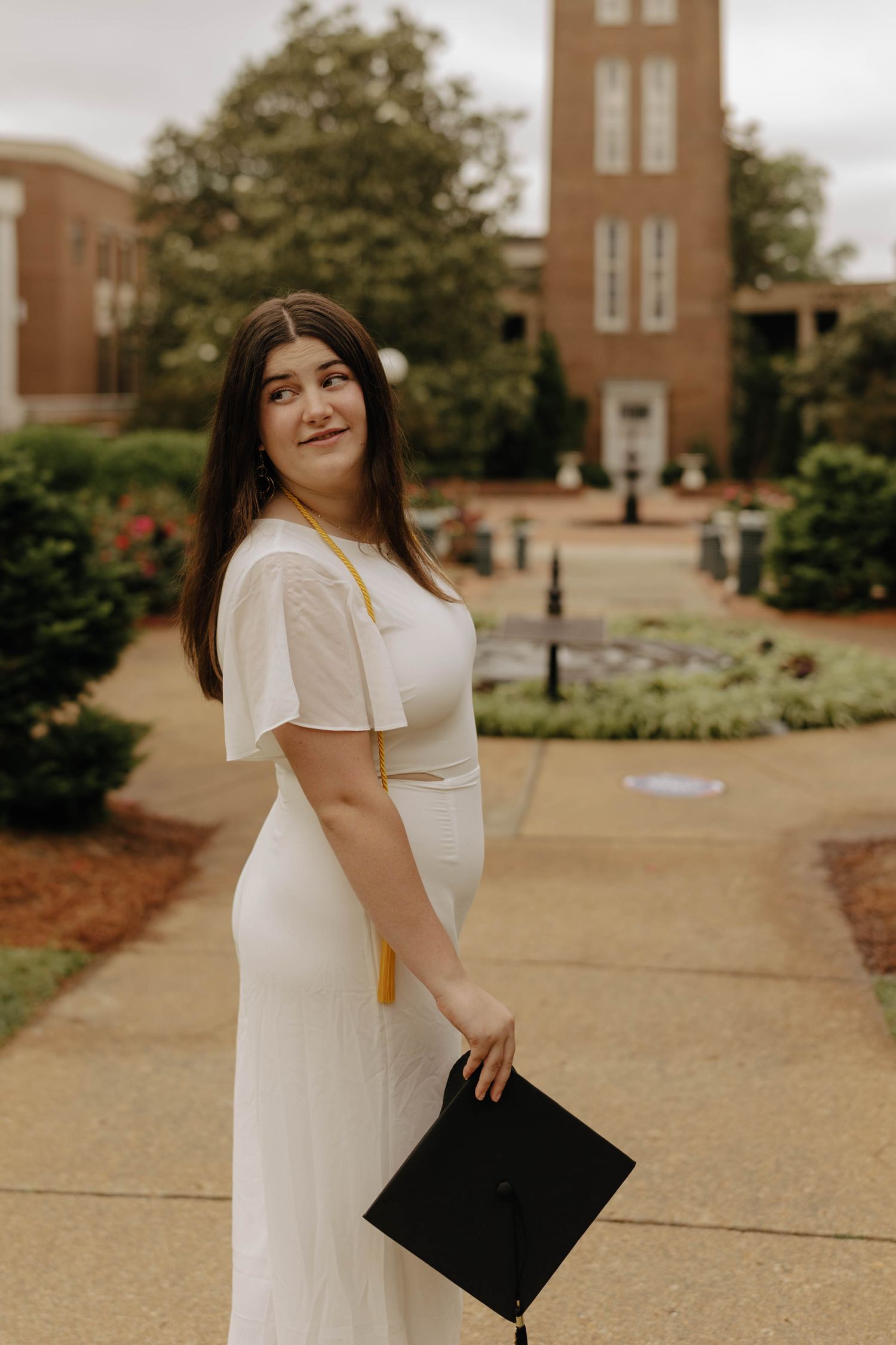 Graduate holding black diploma cover poses on campus walkway near brick buildings.