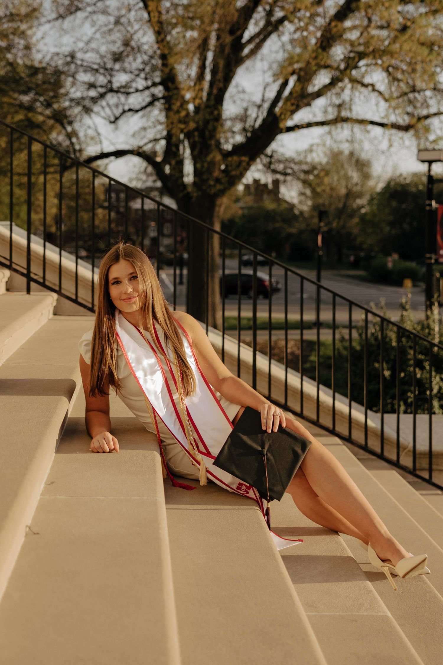 A graduate in academic regalia poses on concrete steps during golden hour.