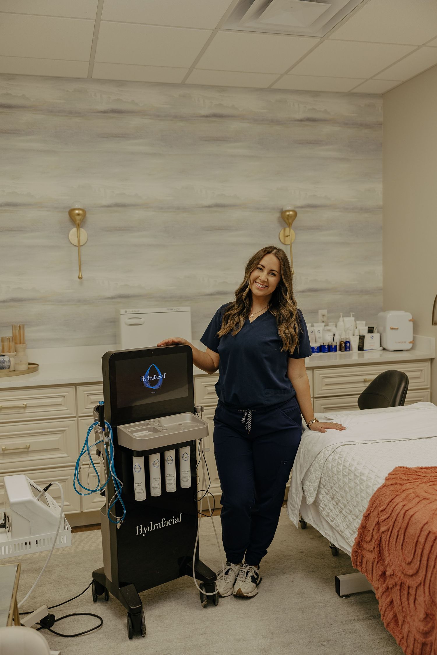 Medical professional in navy scrubs posing in a modern treatment room with bed and equipment.