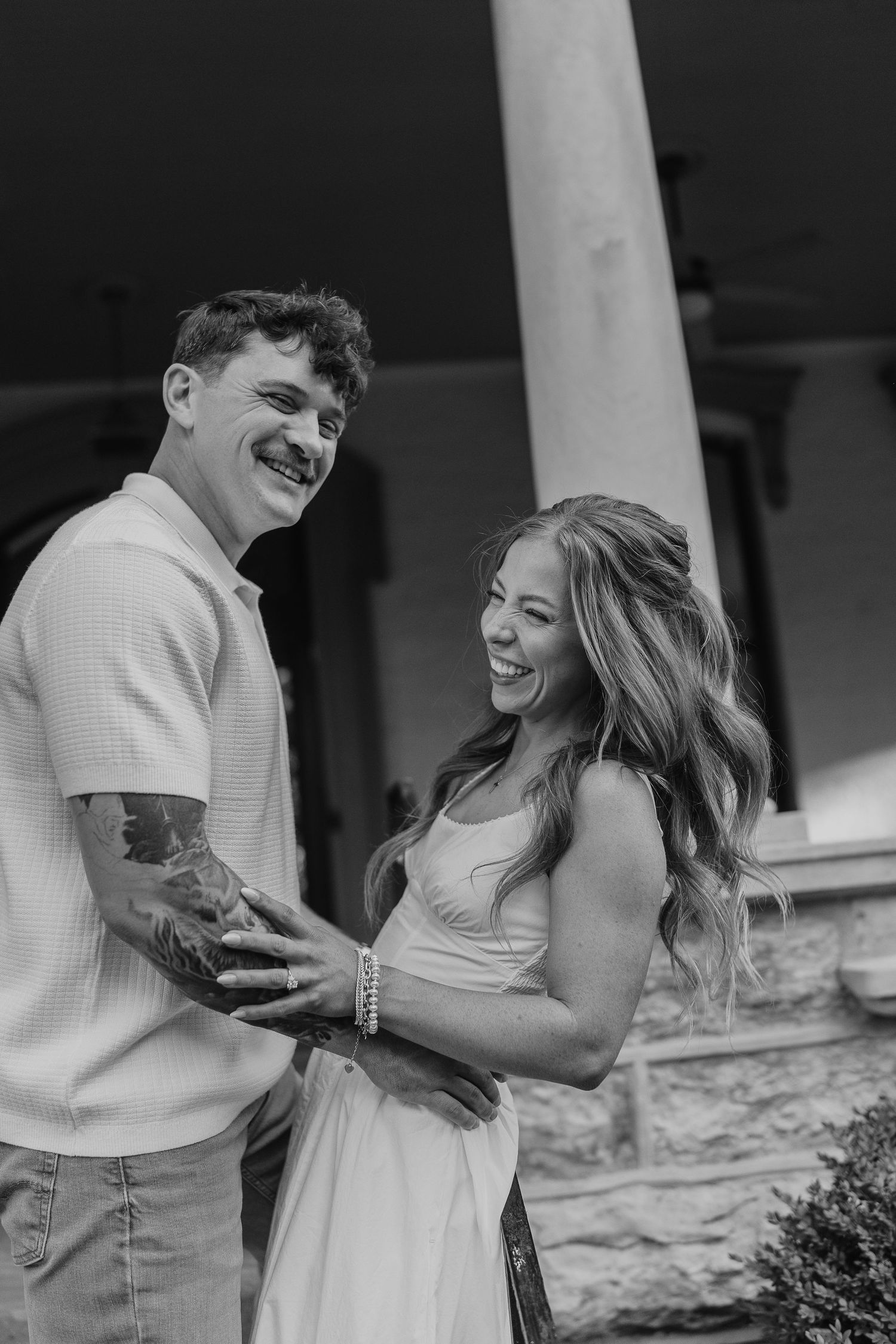 Black and white photo of a smiling couple embracing outdoors near a column, man with tattoos, woman in white dress.
