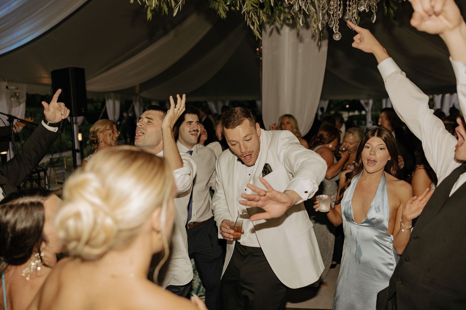 Groom in white jacket celebrates energetically on crowded wedding reception dance floor.