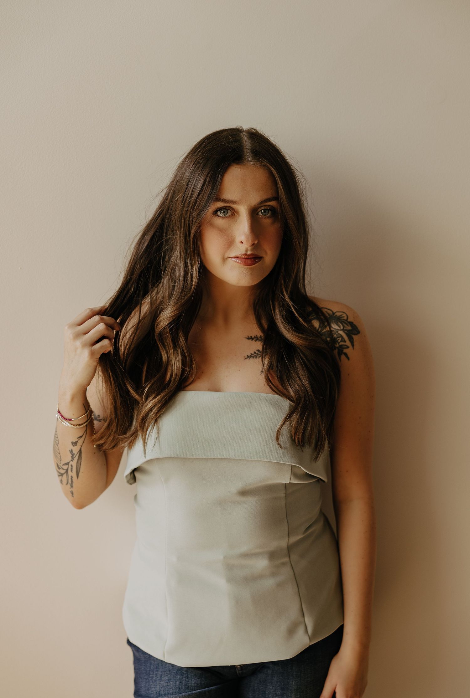 A young woman with long brunette hair wearing a sage strapless top and jeans, with visible tattoos, posing indoors.
