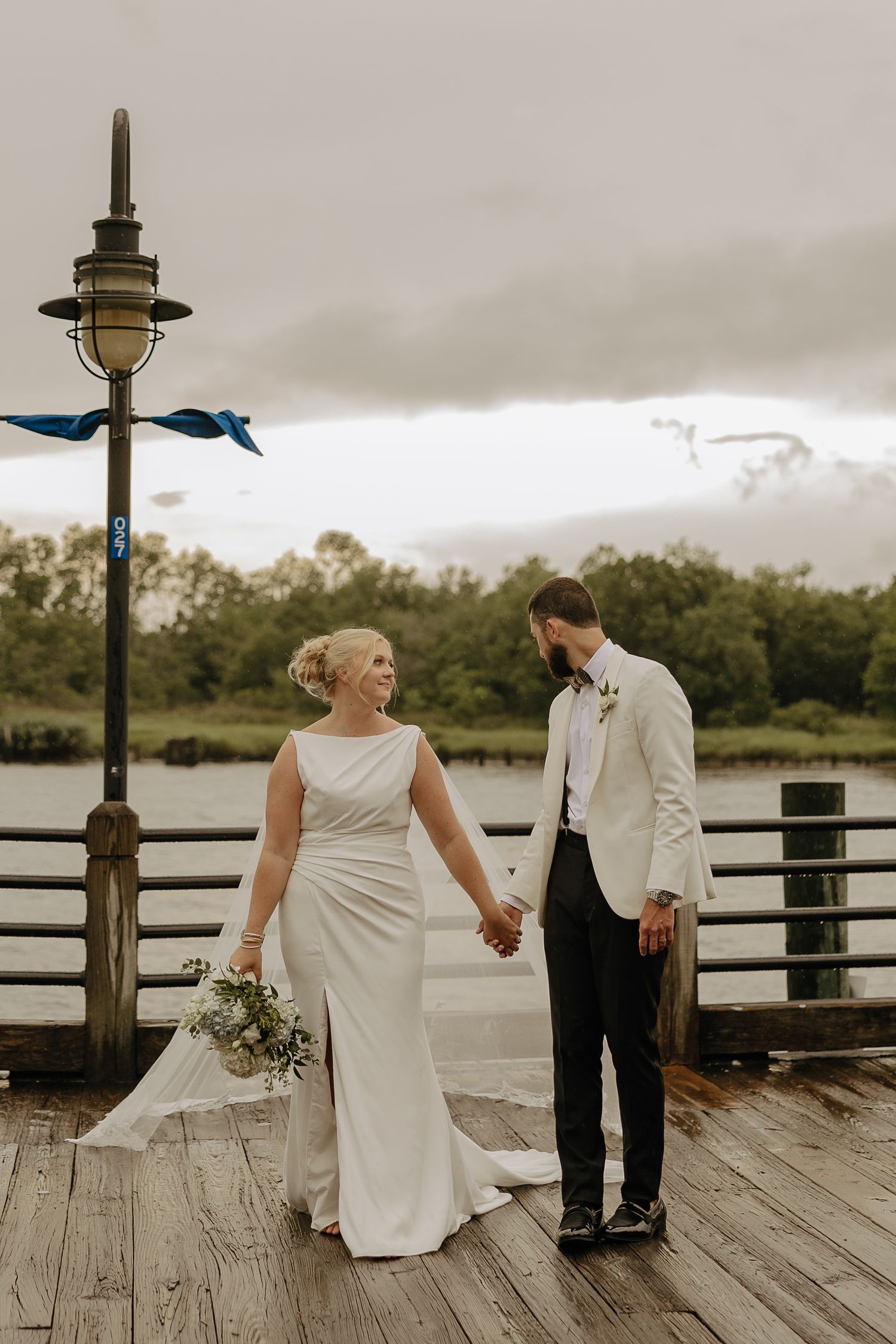 A couple stands on a wooden dock overlooking water on an overcast day.