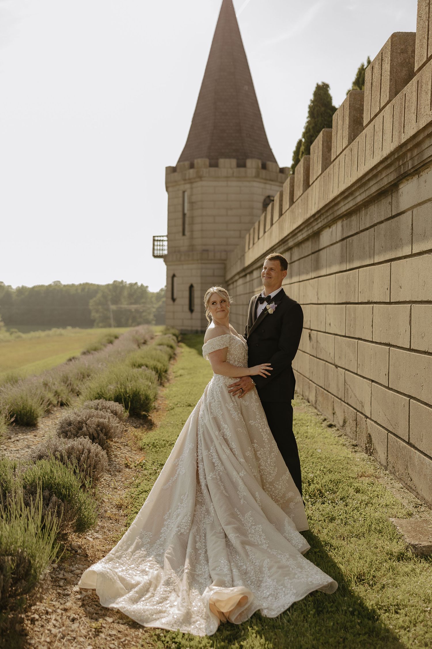A romantic wedding couple poses along a stone wall in an outdoor setting with rolling hills in the background.
