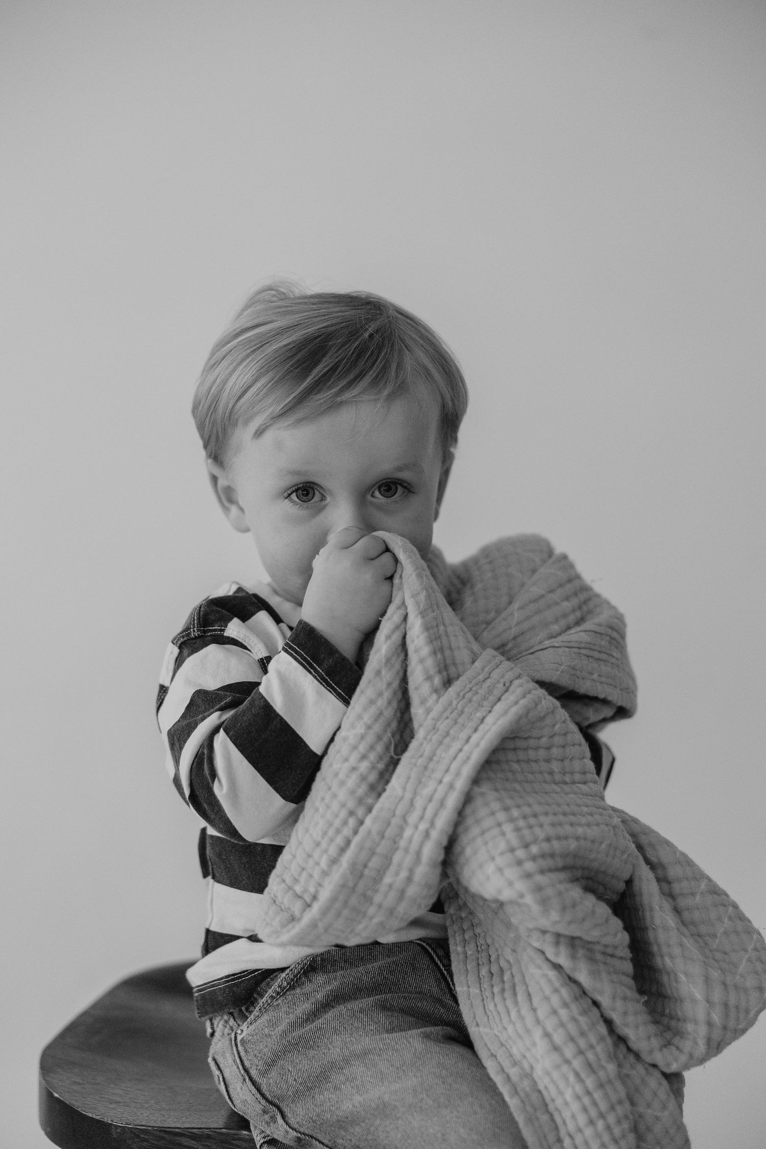 A young child in striped shirt holds a blanket while sitting on wooden stool in black and white portrait.