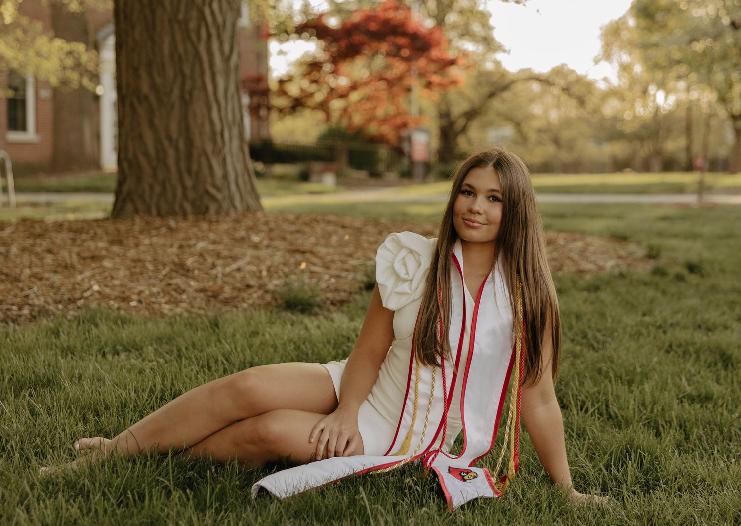 Graduate in white dress with red stoles poses on grass near tree during sunset.