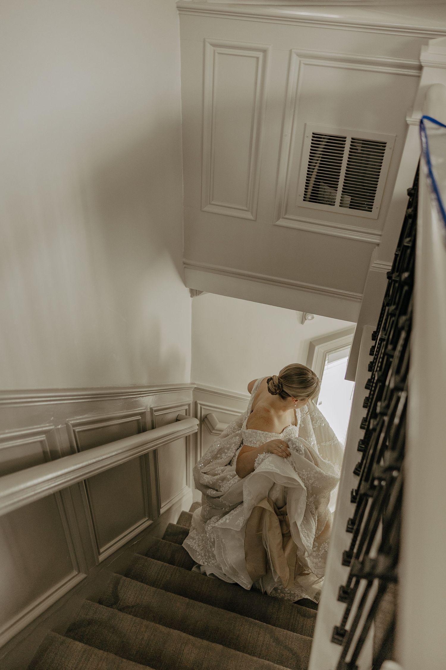 A bride in a flowing white gown descends a classic staircase with wainscoting.