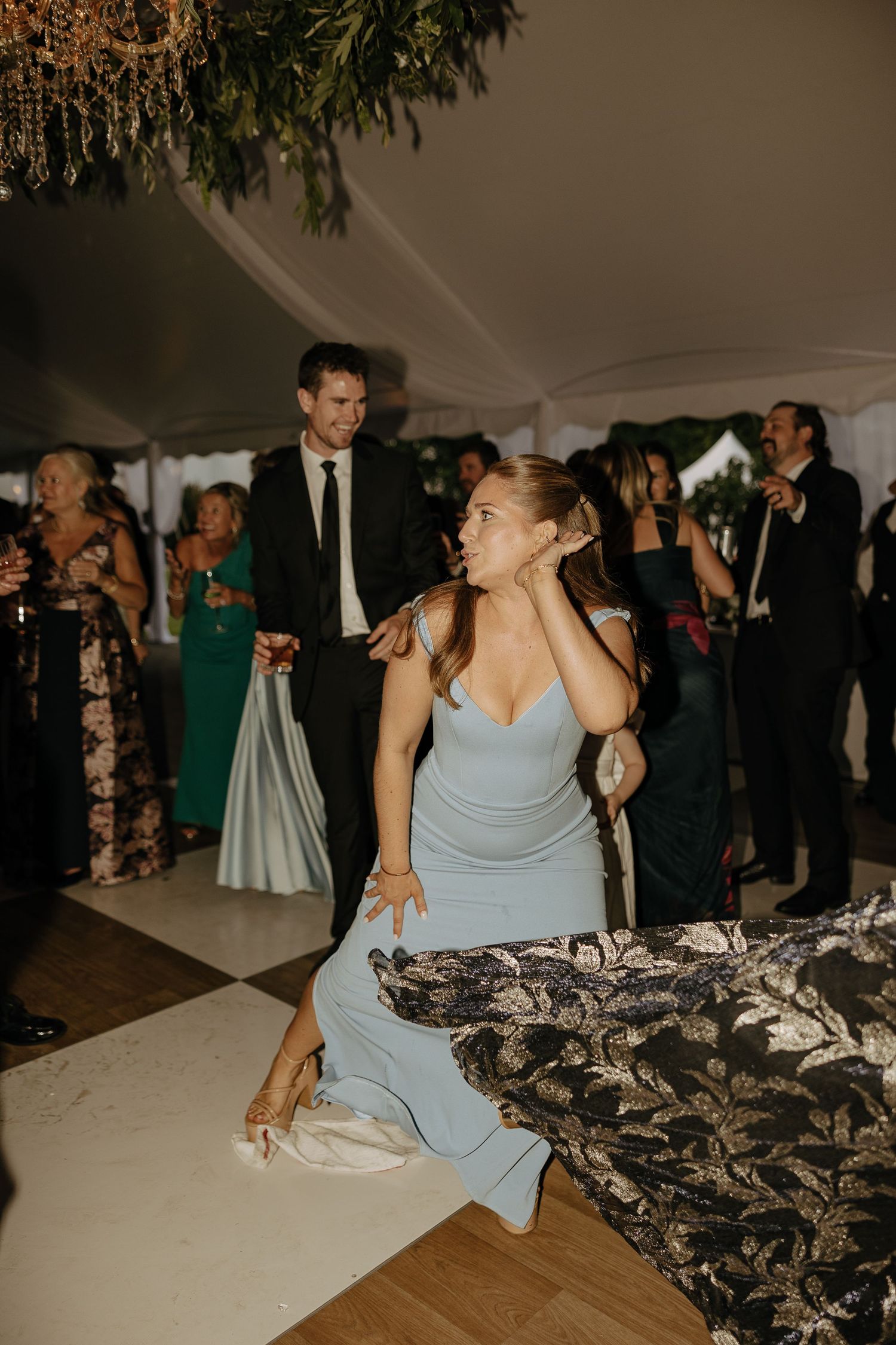 Bridesmaid in light blue gown sitting on dance floor during lively outdoor tented wedding reception.