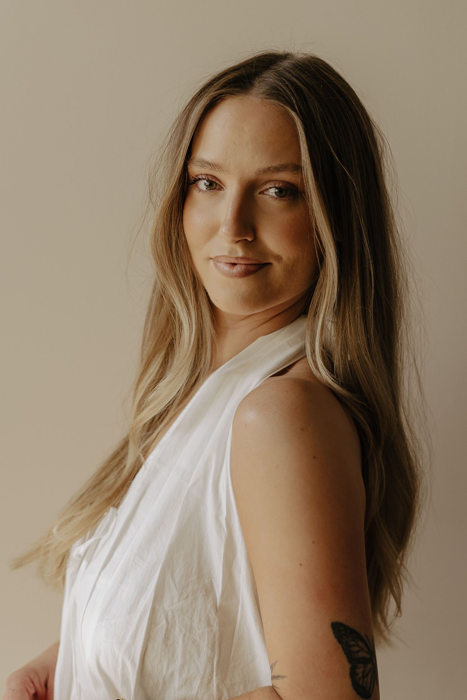 Young woman with long blonde hair wearing a white sleeveless top, showing a tattoo on her arm, posing against a beige background.