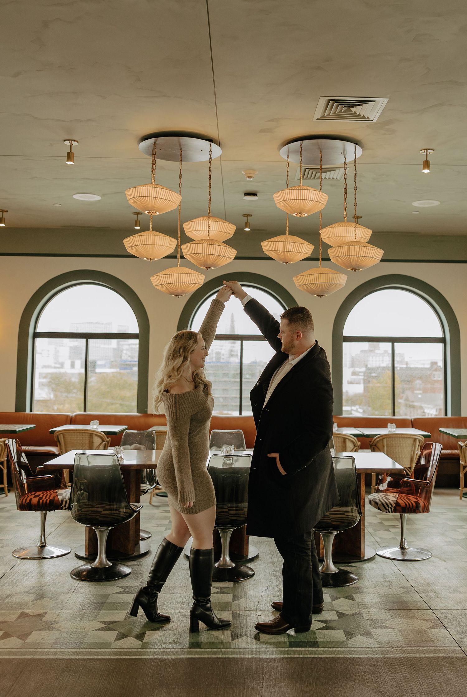 Two people dance together in an empty dining room with modern decor and large arched windows.