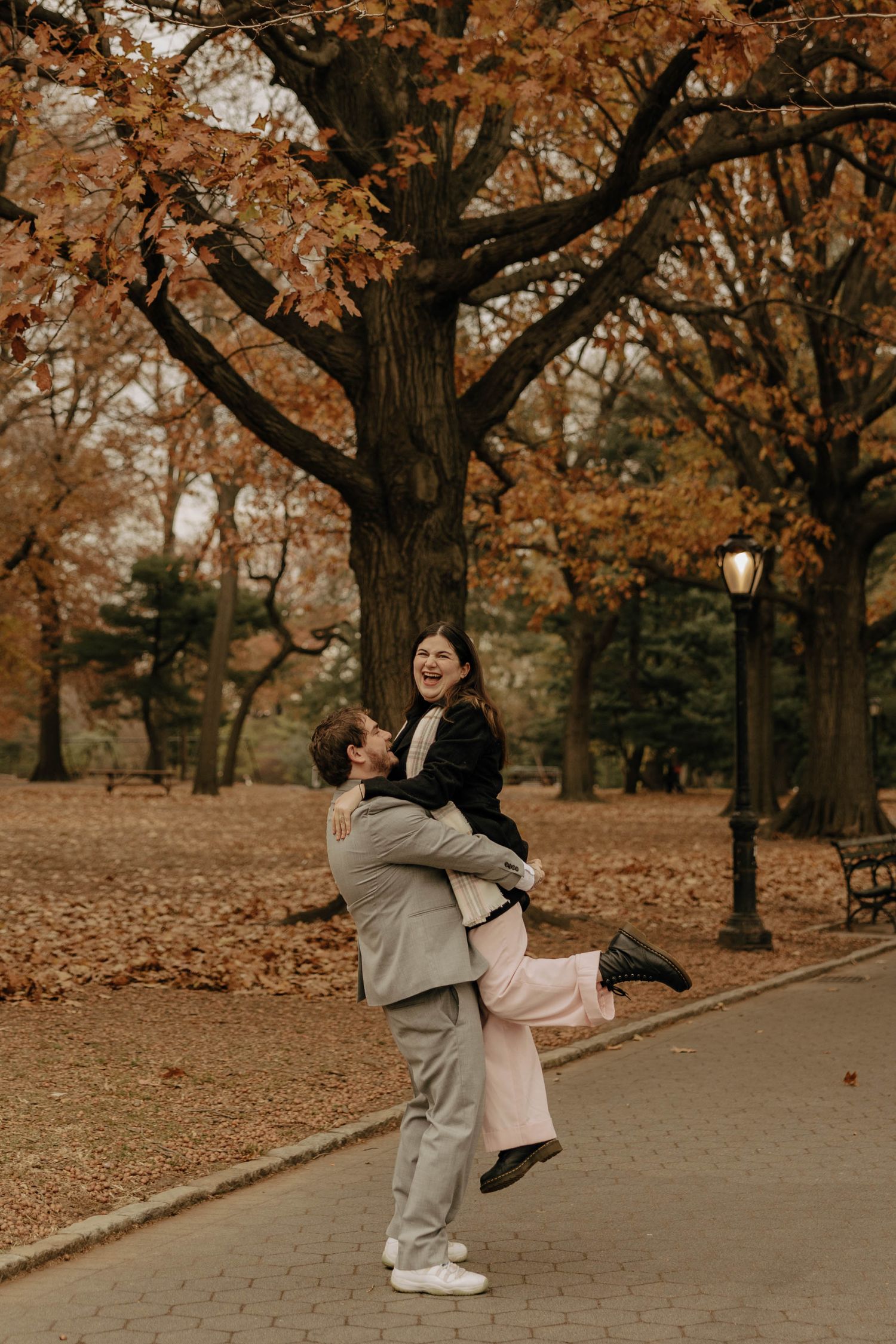 A joyful couple embraces and laughs together on a path surrounded by fall foliage in the park.