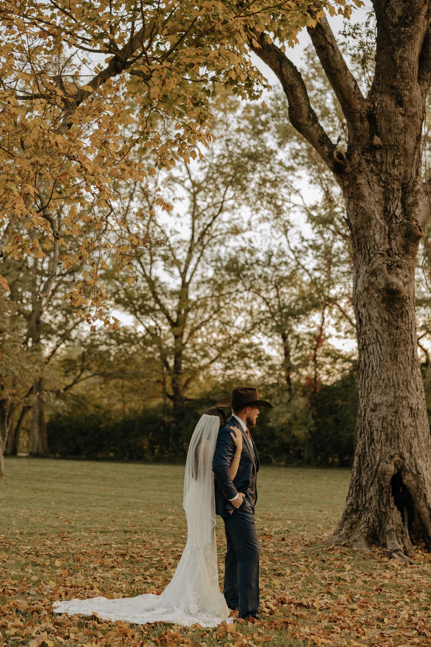 A couple in wedding attire poses for photos among autumn trees with fallen leaves on the ground.