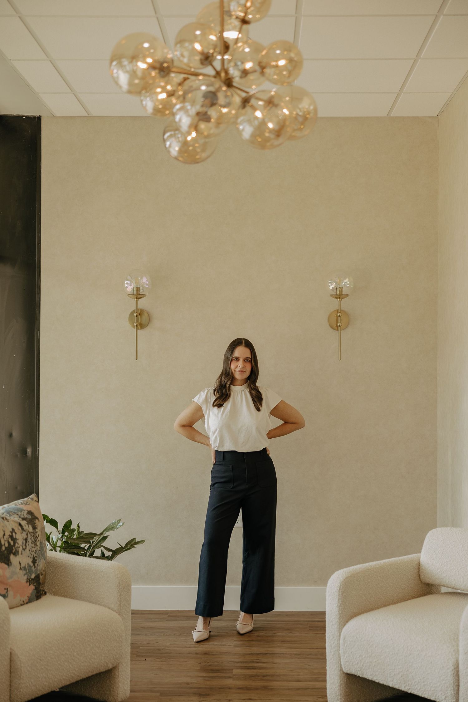 Full length portrait in elegant white top and black pants under modern chandelier in studio setting.