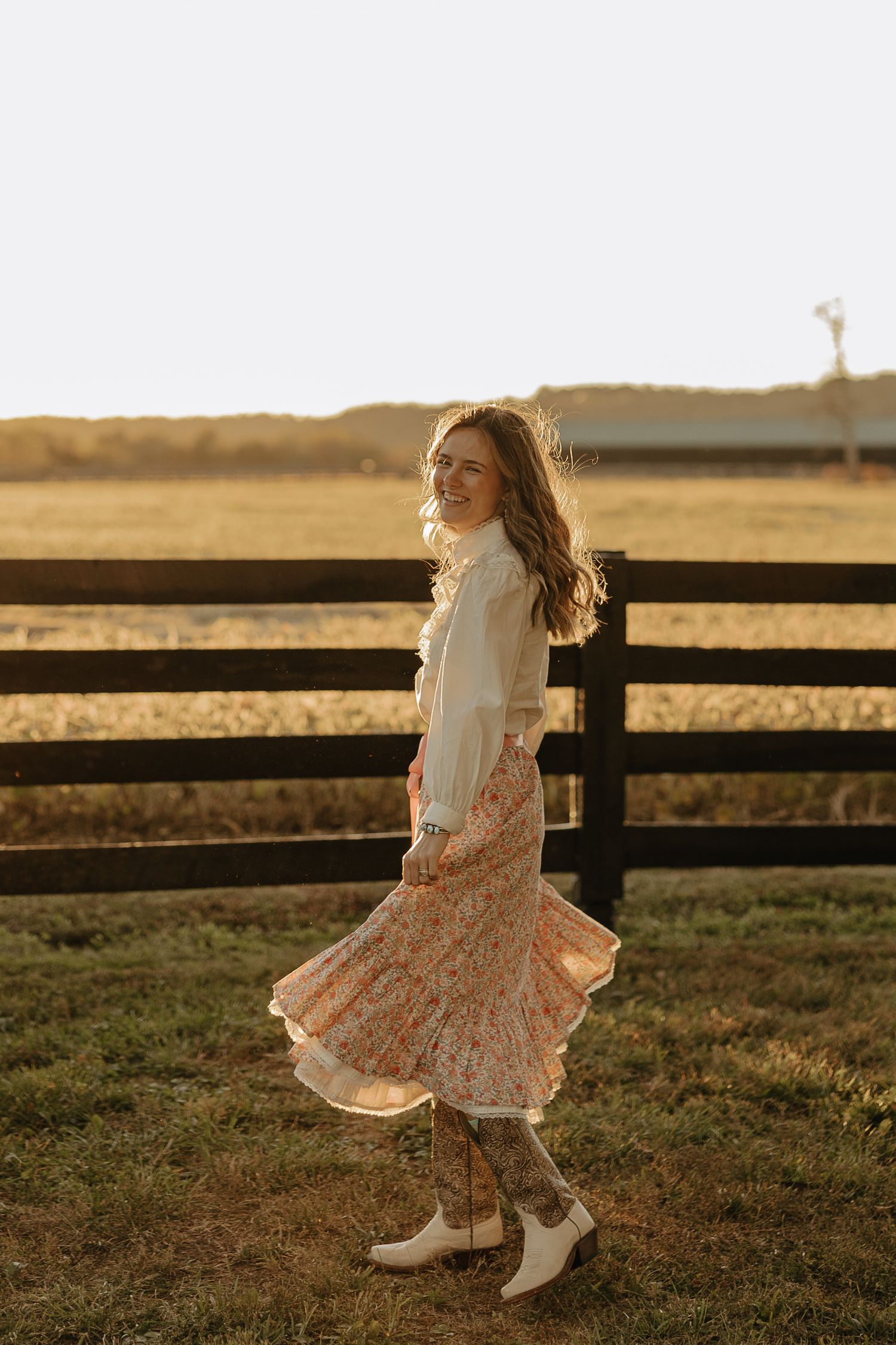 Person twirling in a floral dress and white boots at sunset near a wooden fence.