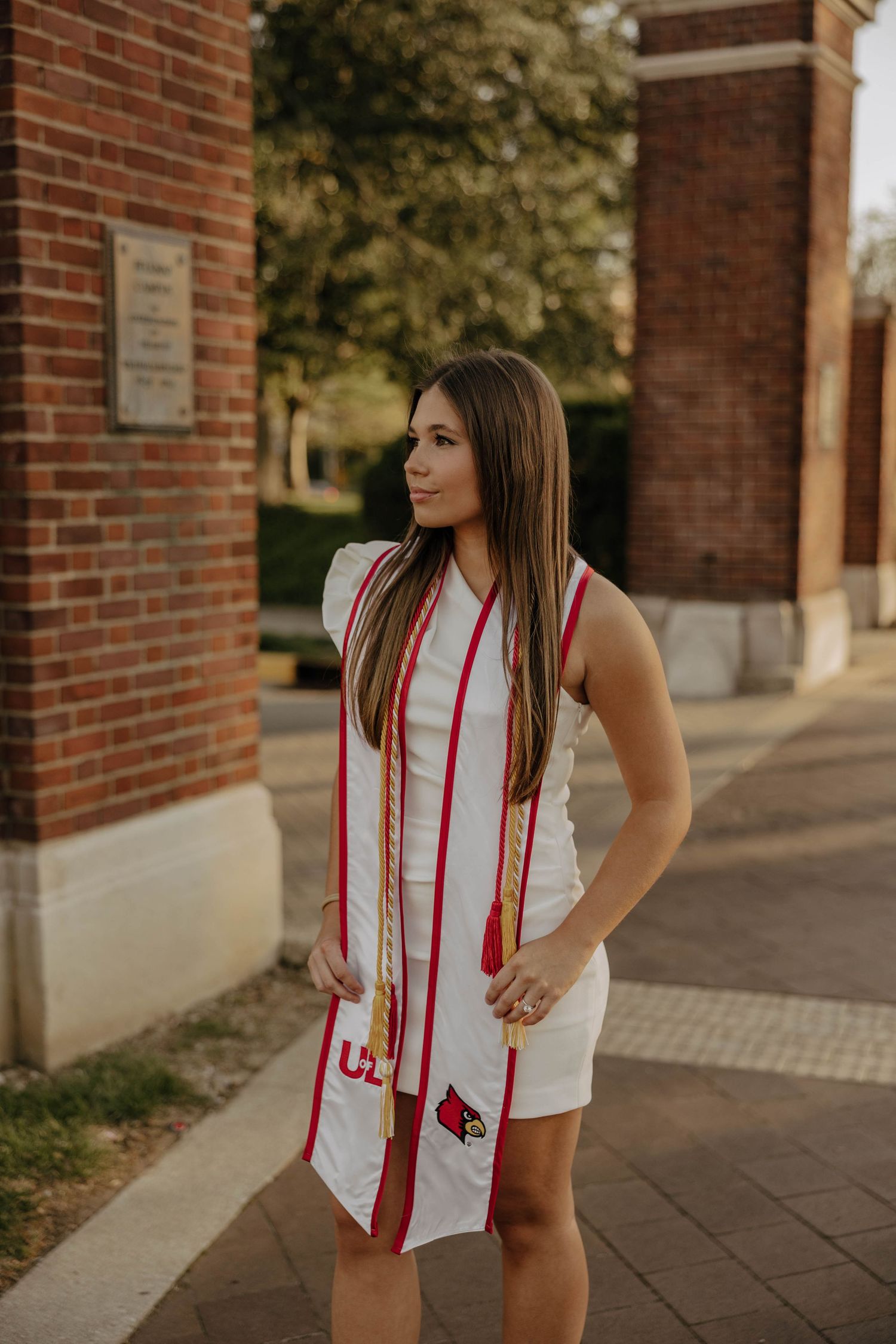 Graduate in white dress posing with red graduation stole near brick columns.
