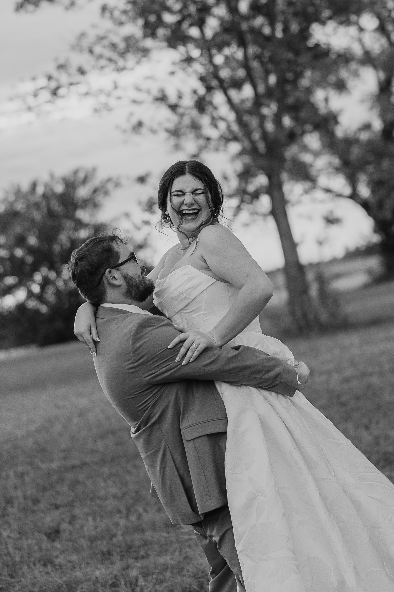 A joyful couple shares a playful embrace outdoors in black and white as the groom lifts his bride during their wedding photoshoot.