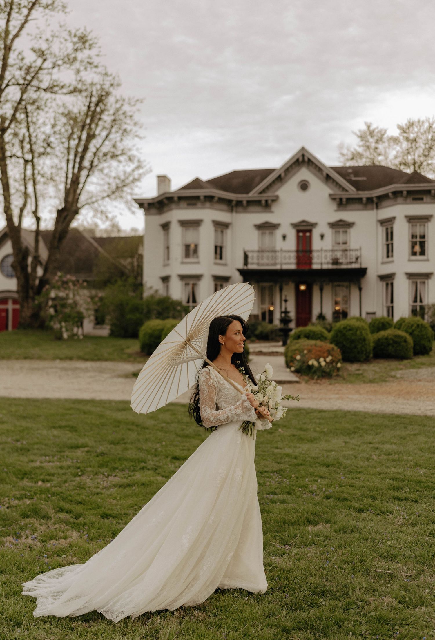 A bride in a flowing white dress with puff sleeves twirls gracefully on a grassy lawn in front of a white historic building.