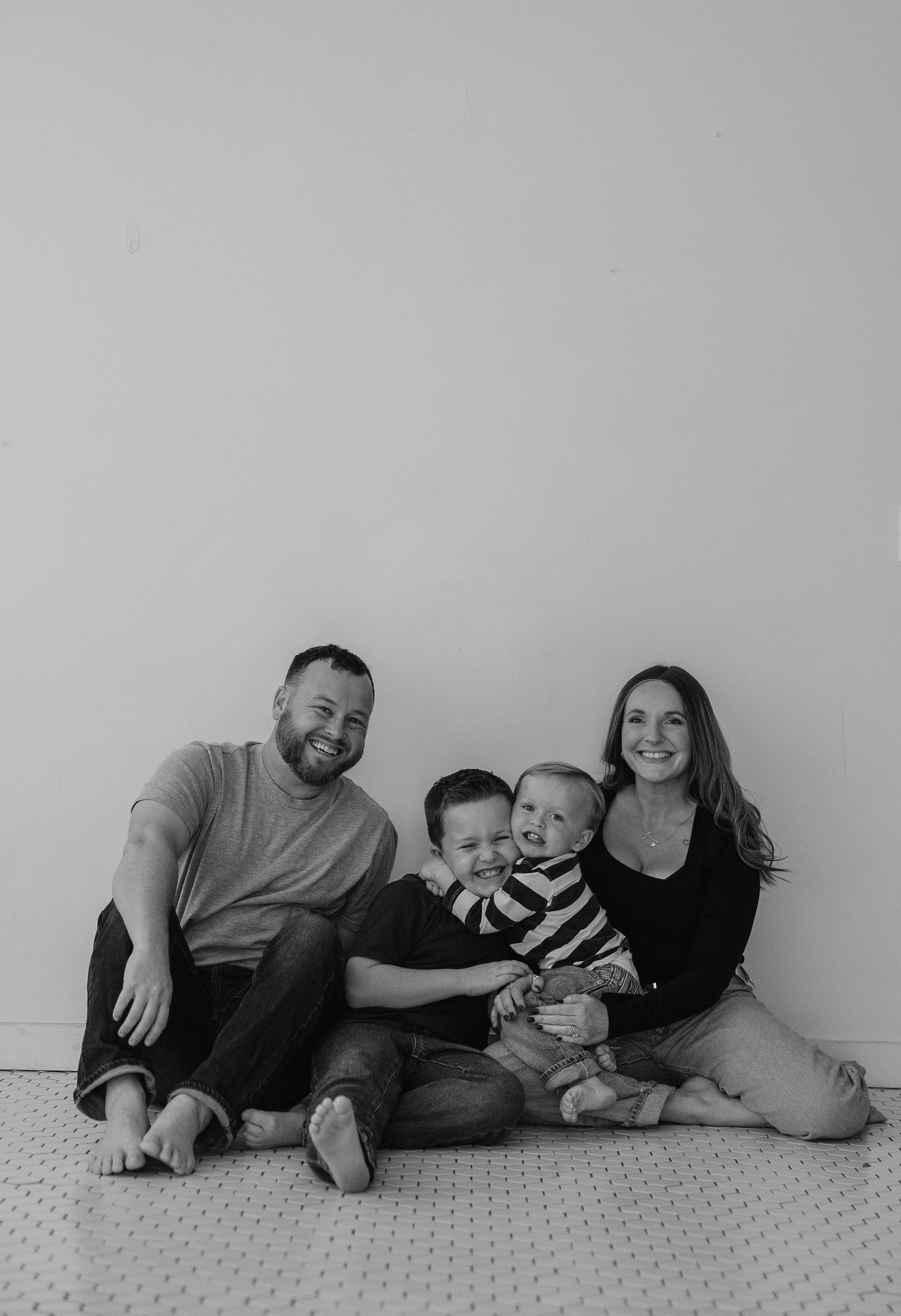 A family of four sits together on a carpeted floor against a plain gray wall in a black and white photo.