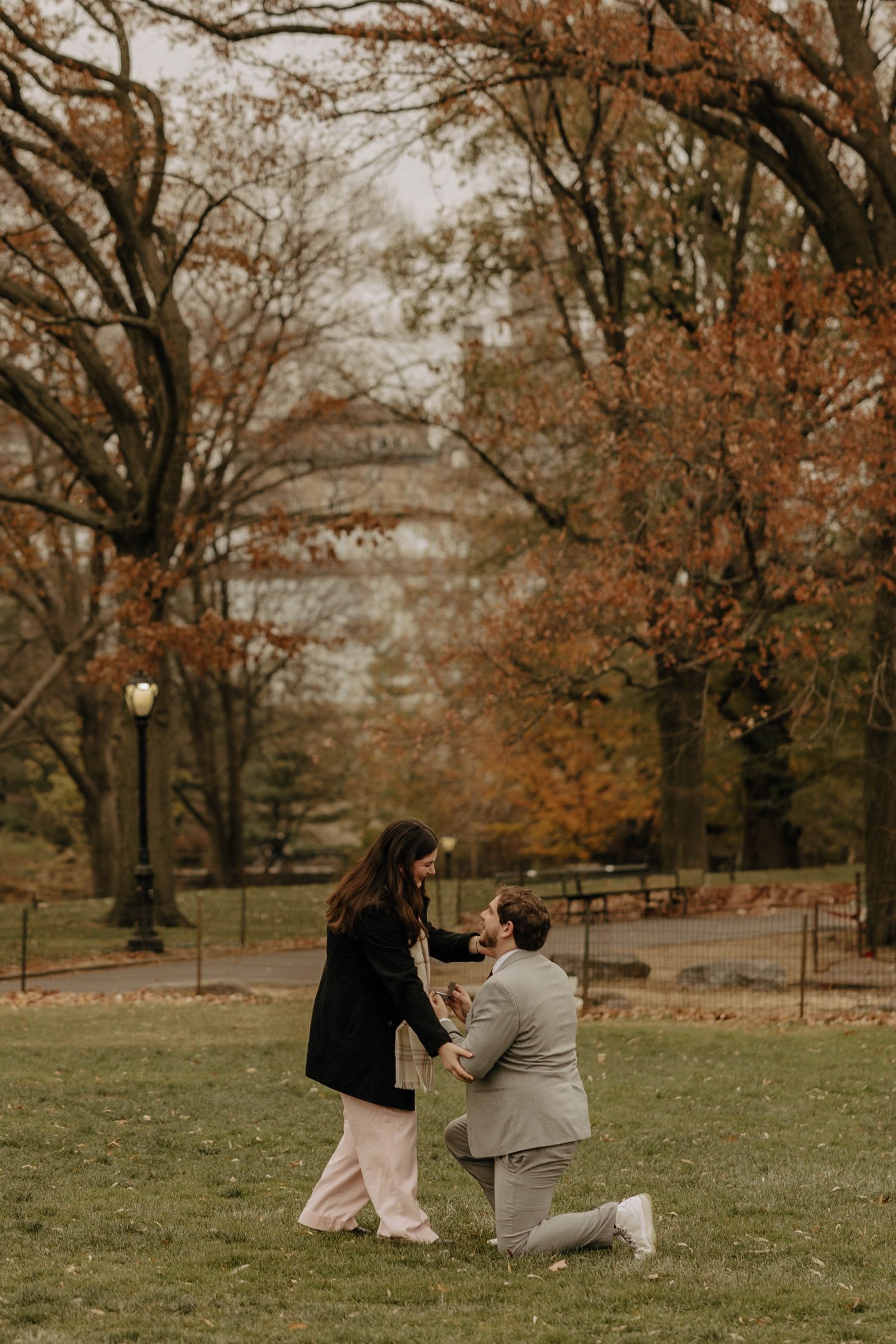 A couple shares a romantic moment in a park on an autumn day with fallen leaves and bare trees in the background.