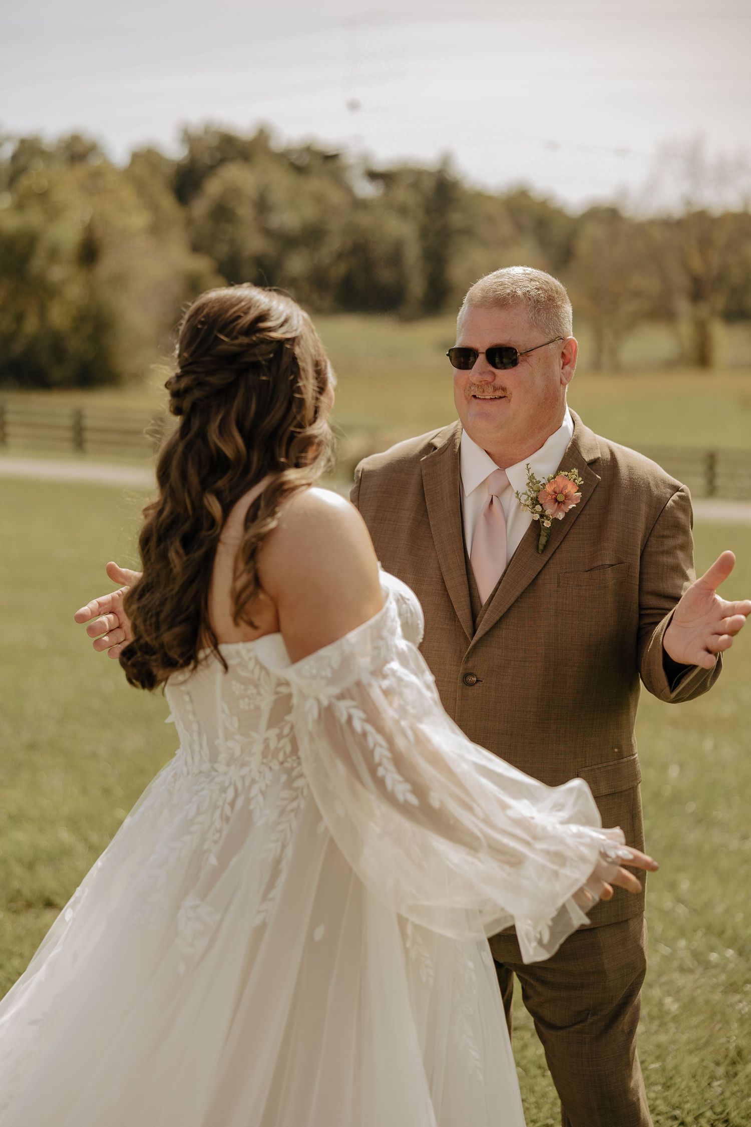 A bride in a white lace dress dances with someone wearing a brown suit in an outdoor setting.