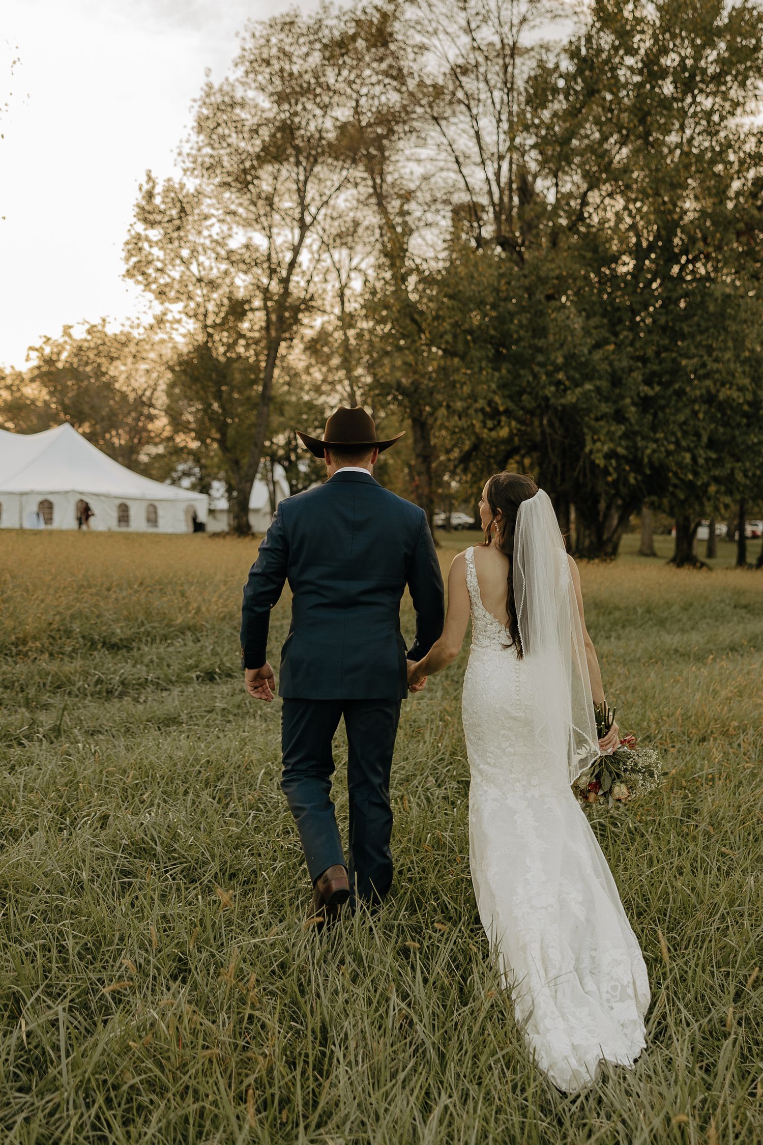 A newlywed couple walks hand in hand through a sunlit meadow.