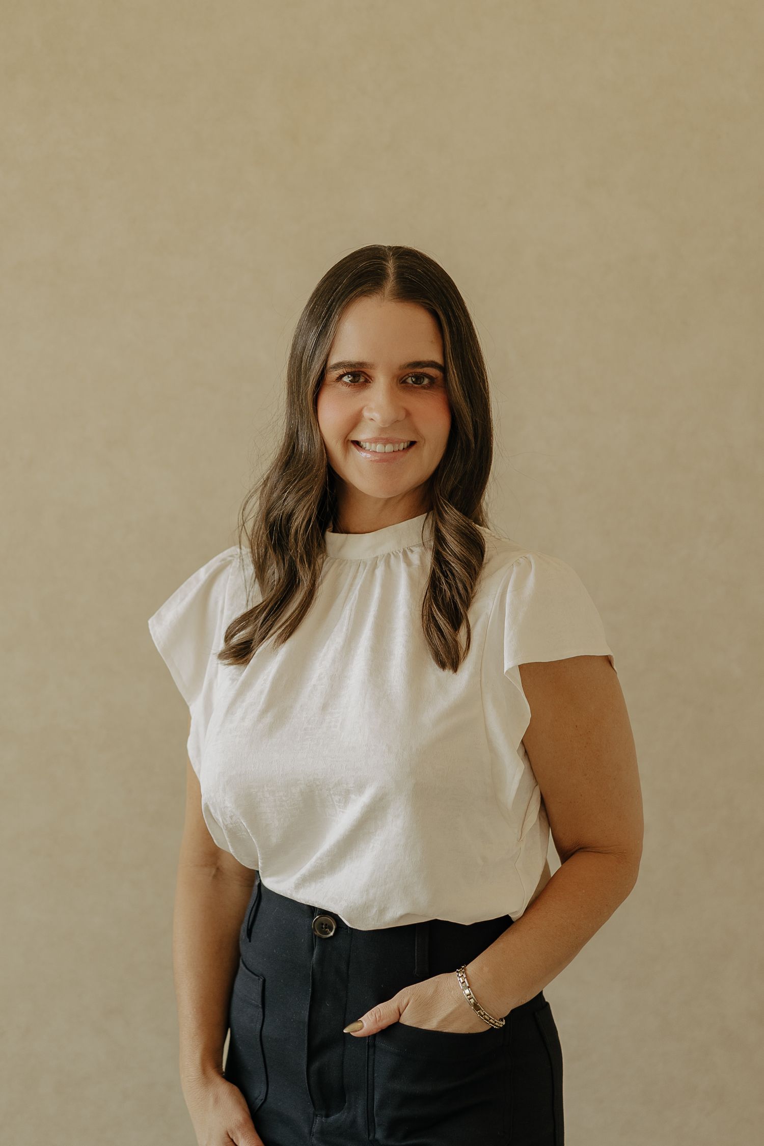Professional headshot in white blouse and dark pants against neutral backdrop.