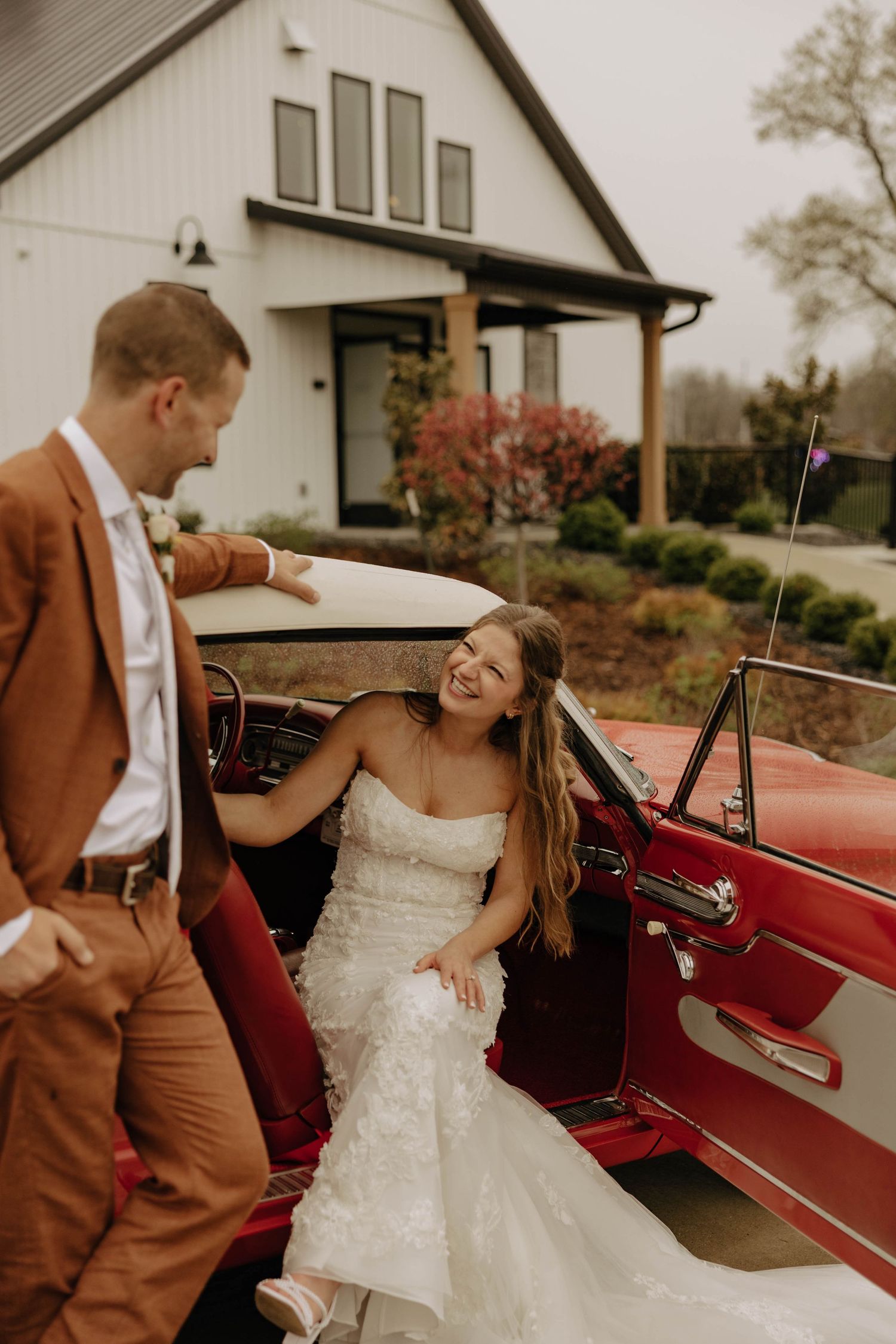 Couple shares joyful moment near red vintage car outside white building.
