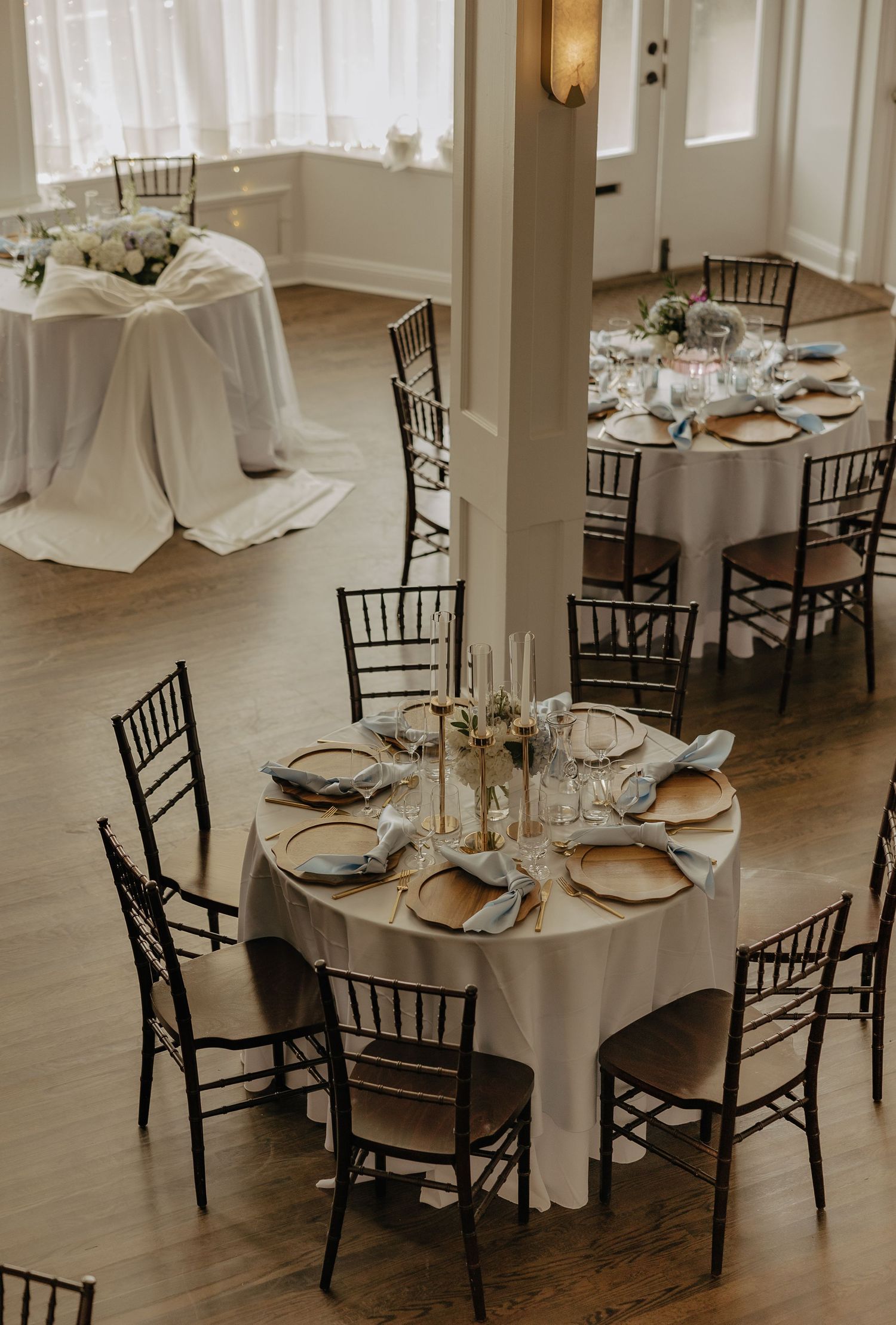 Elegant wedding reception setup with white tablecloths and black chiavari chairs in a bright room.