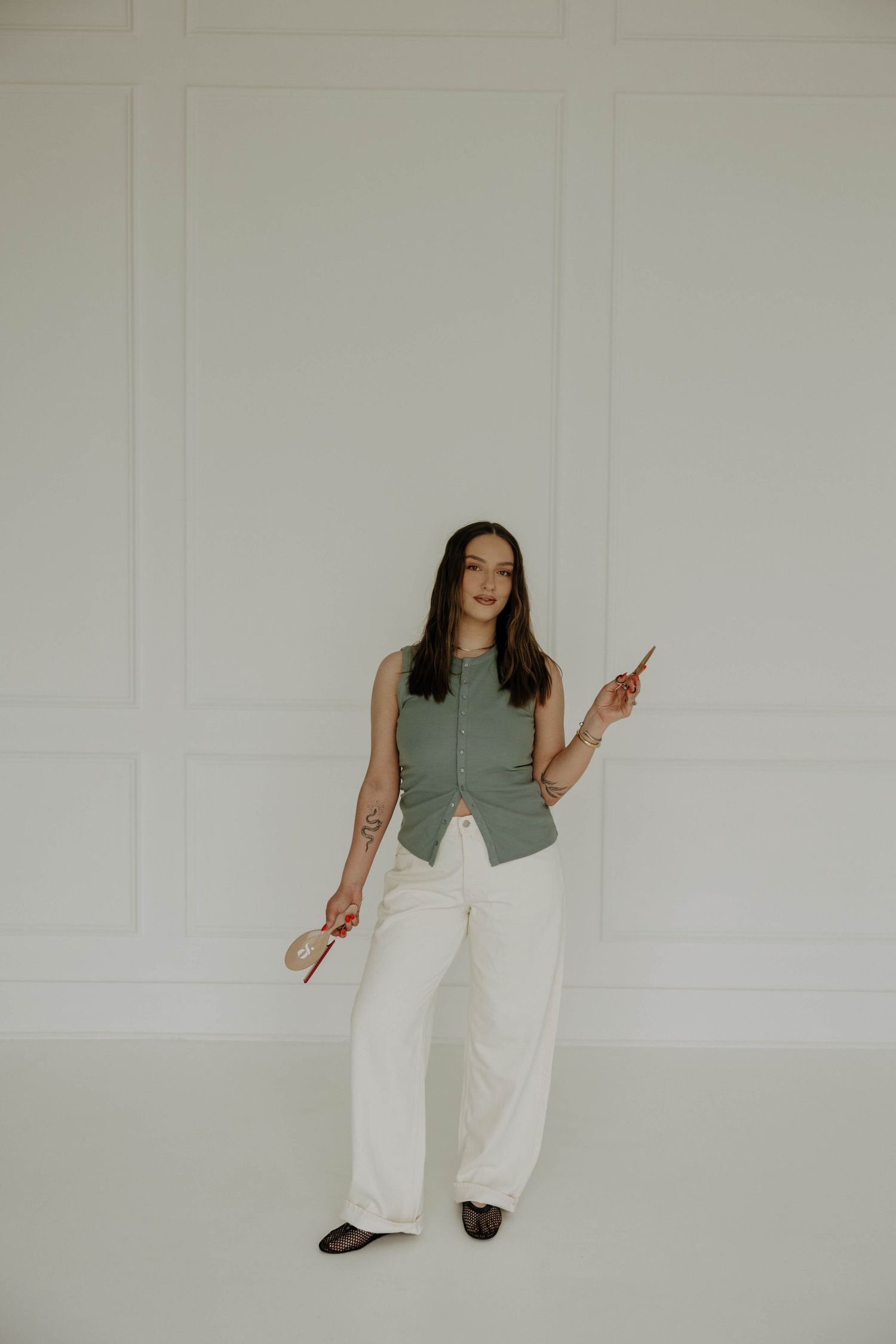 A person in a sage green button-up top and white wide-leg pants poses against a white wall.