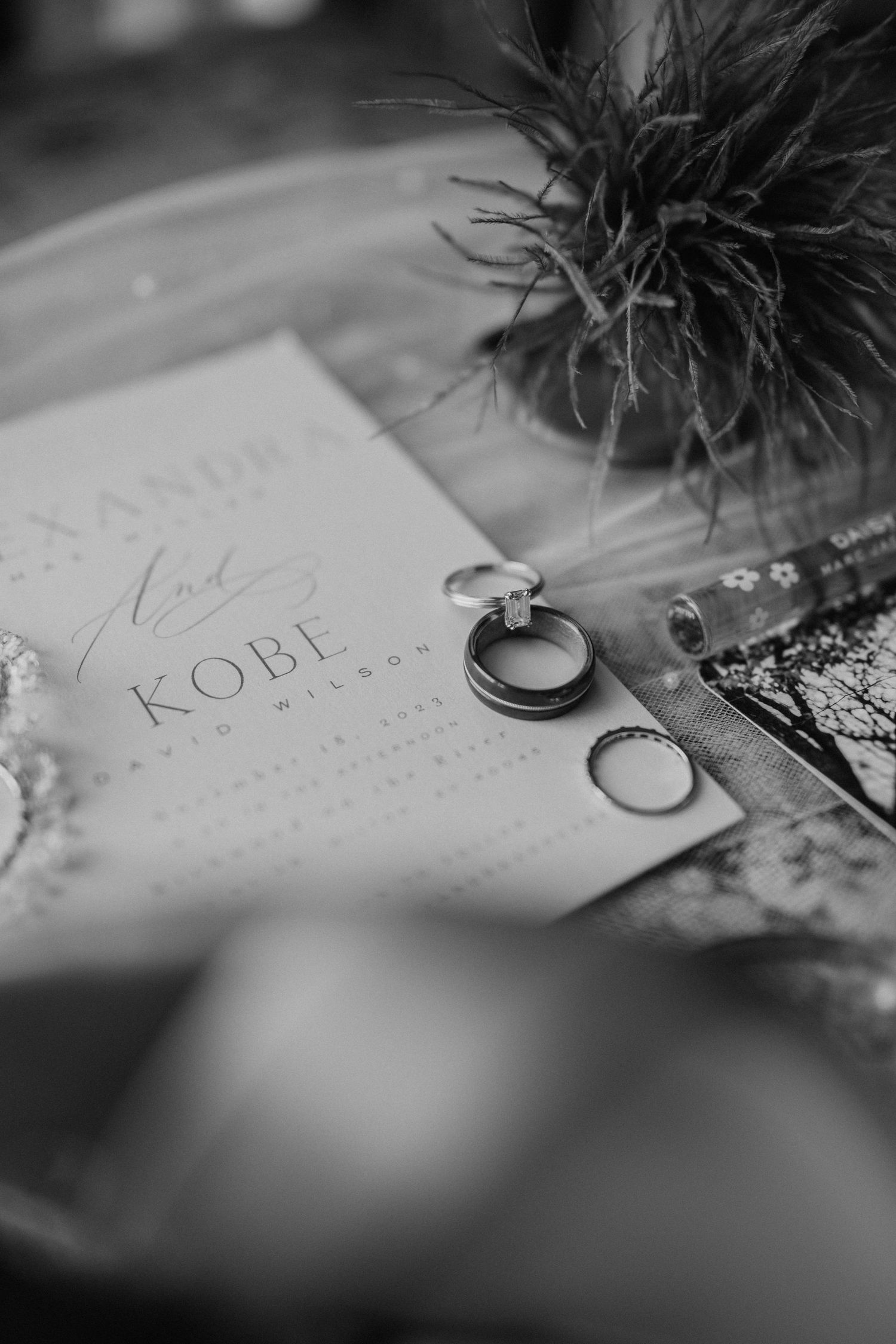 Black and white wedding detail photo showing rings and invitation with Kobe text alongside a decorative plant.