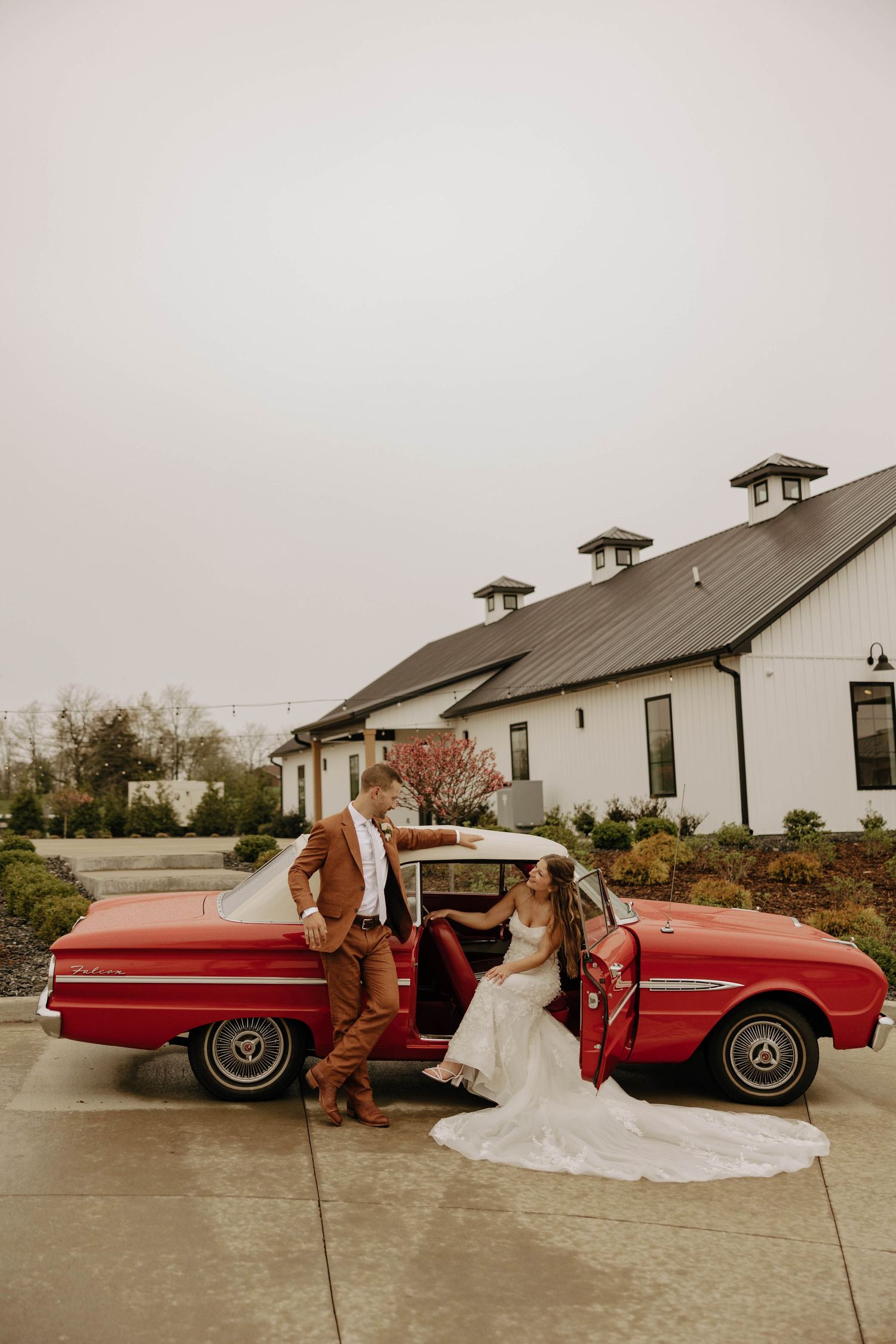 A vintage red convertible car parked outside white buildings with figures posing in wedding attire in a photo series.