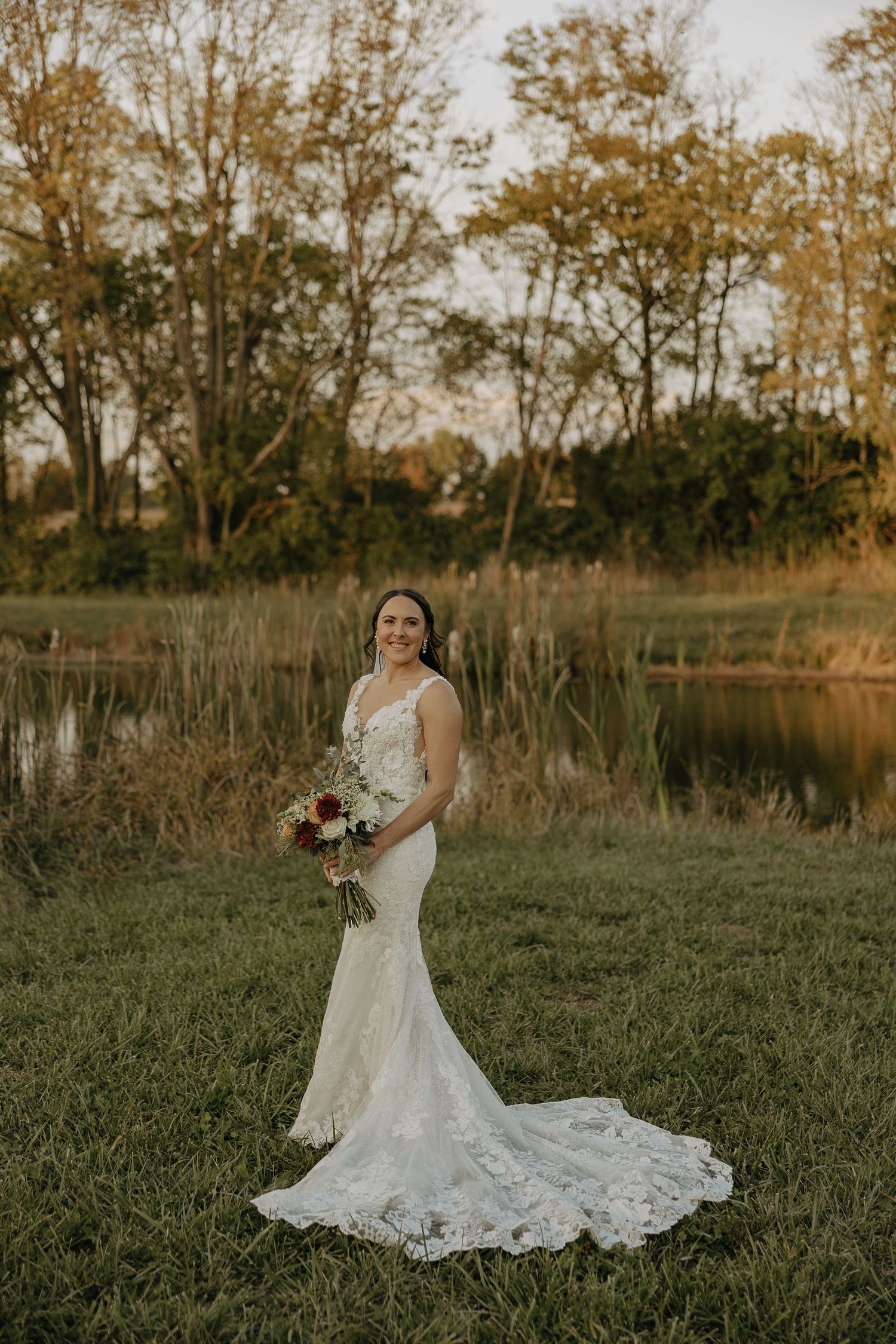 A bride in a lace wedding gown poses by a pond during golden hour.