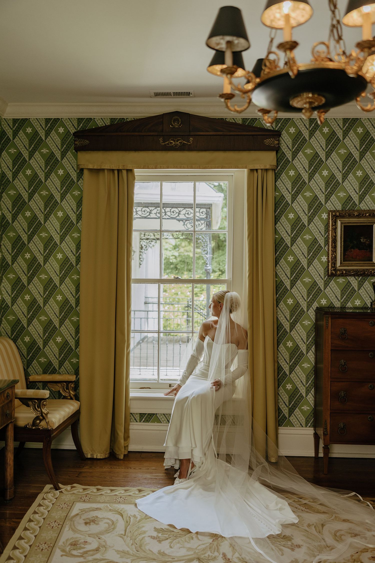 Bride in long-sleeved gown and veil sits by window in green wallpapered room on wedding day.
