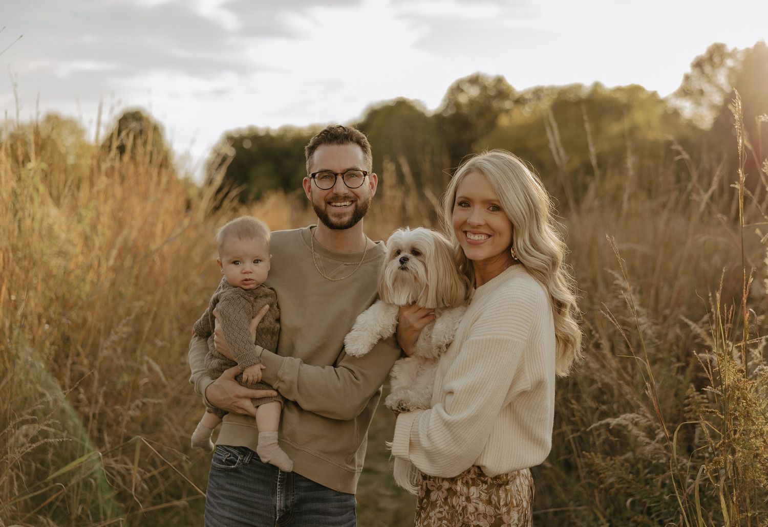 A family poses together with their small white dog in tall autumn grass during golden hour.