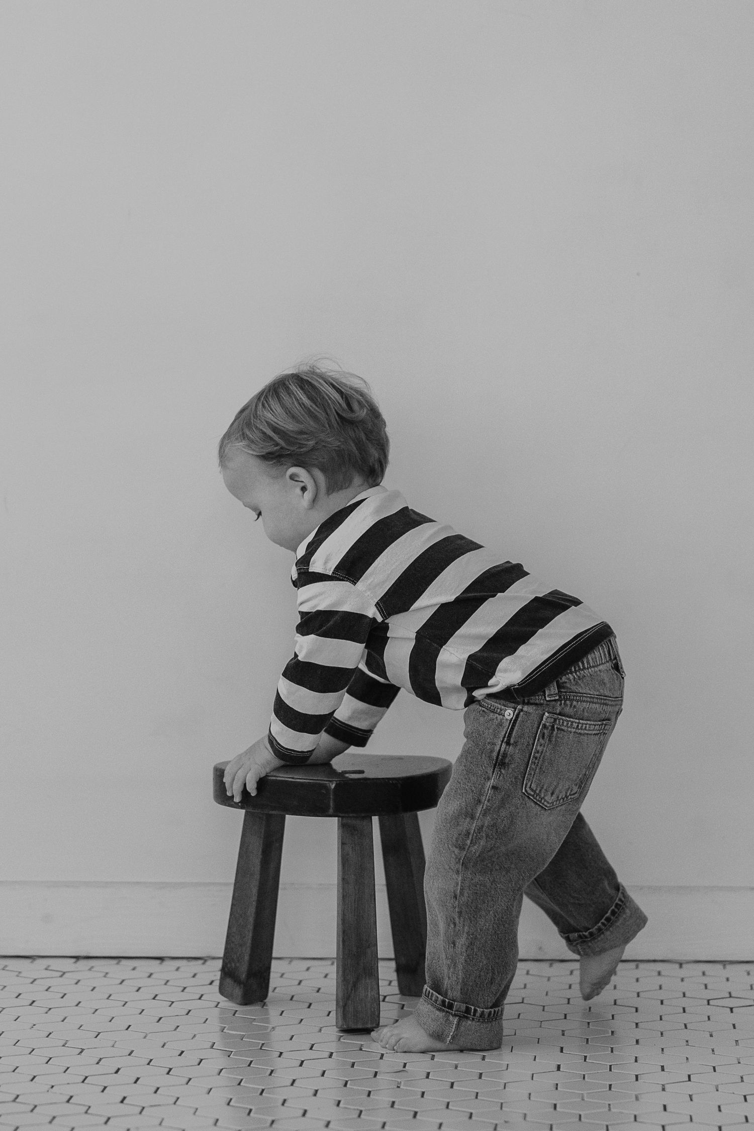 Side view of small child in striped shirt climbing wooden stool in black and white photo.