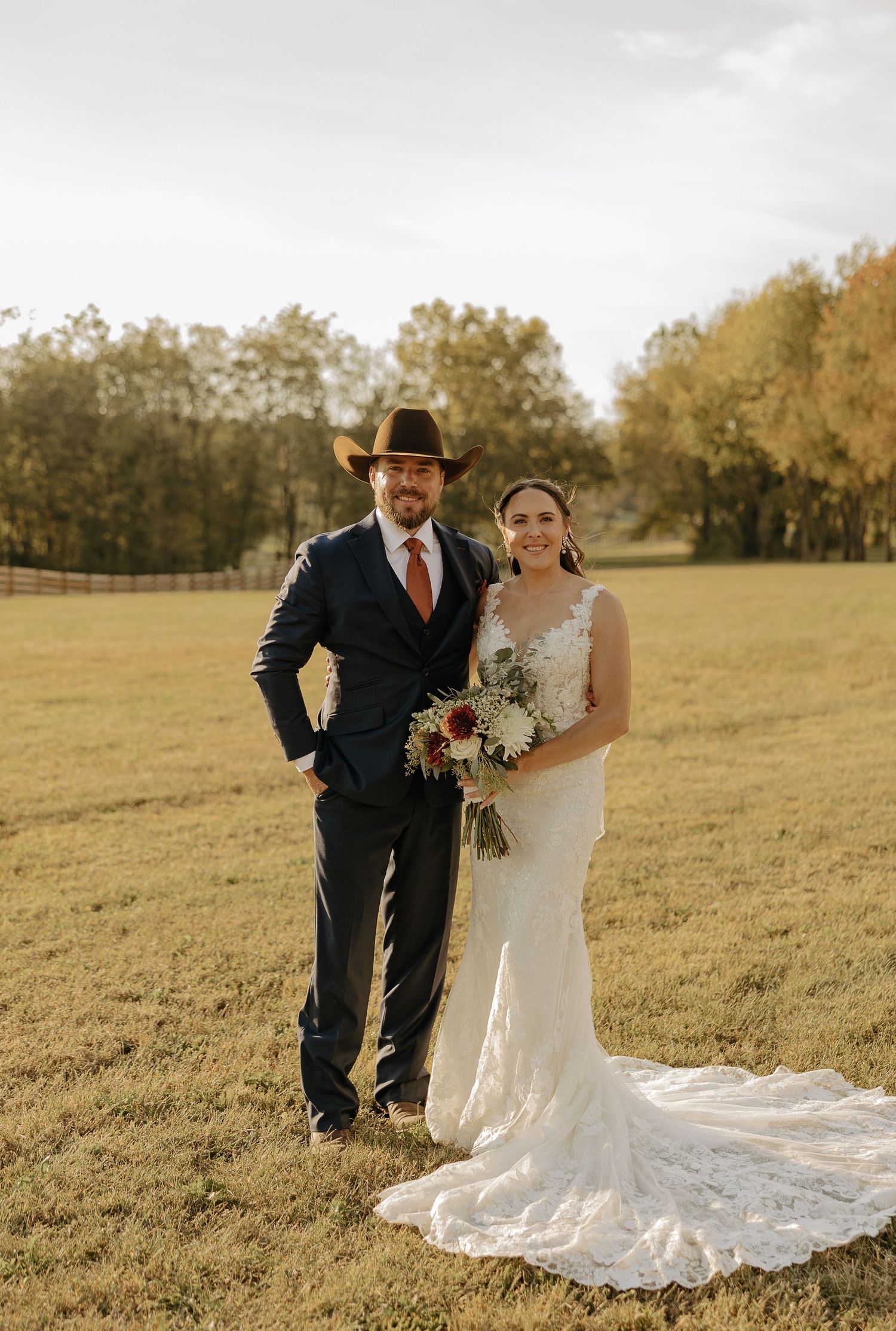 A couple poses in a golden field during sunset with the bride wearing a flowing lace gown.