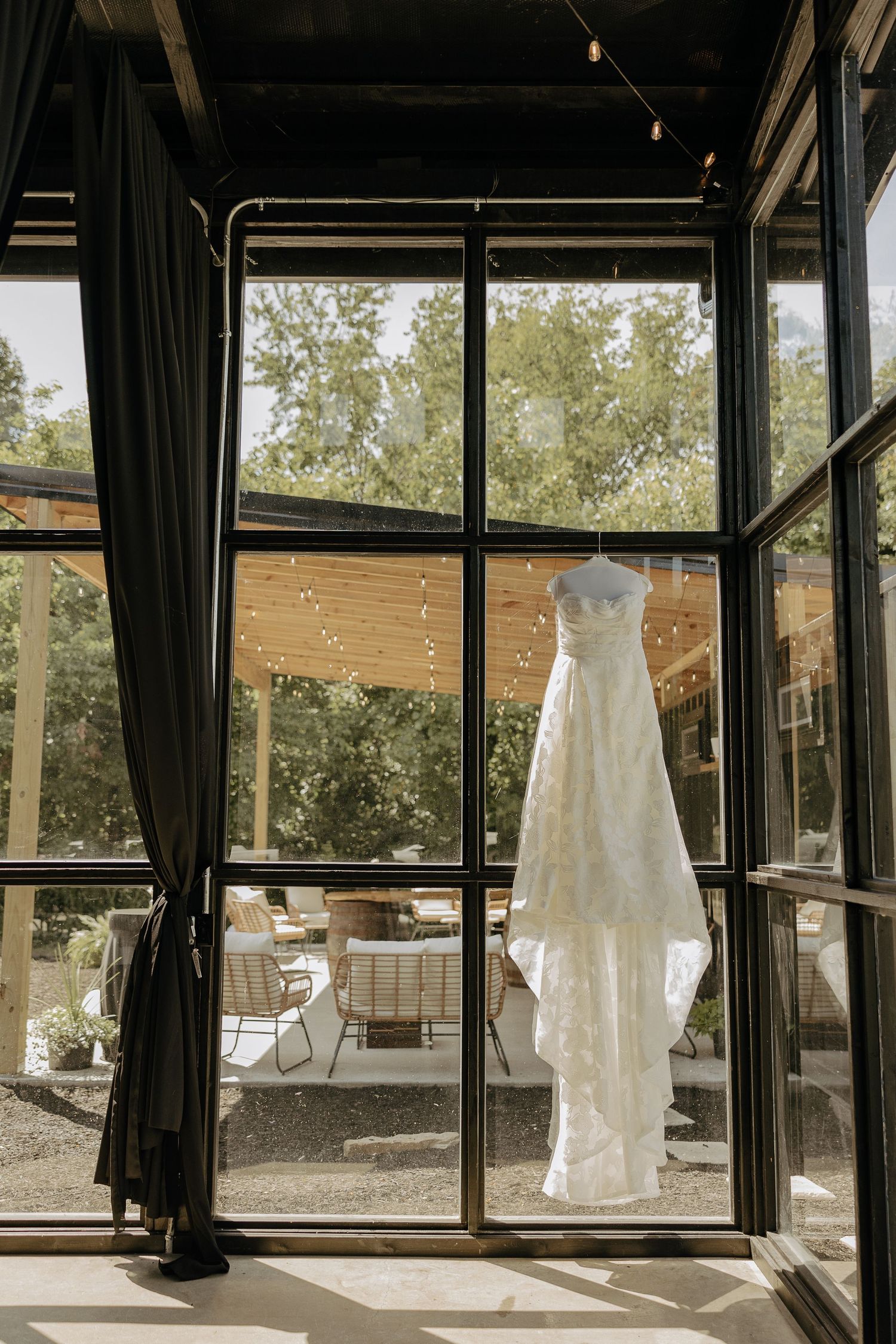 Wedding dress displayed in sunlit window of modern venue with dark curtains and outdoor patio view.