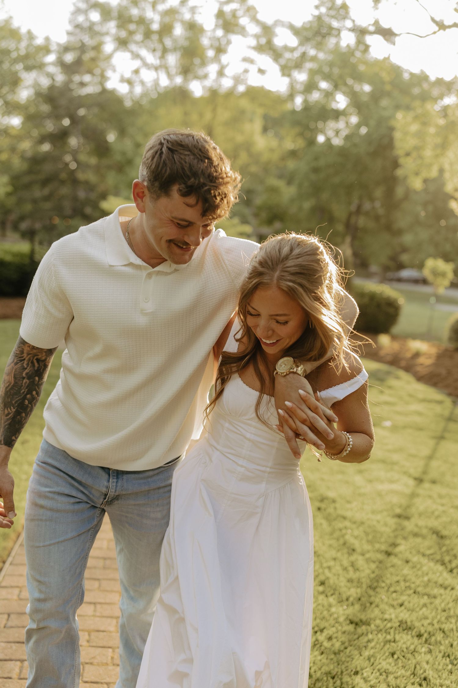 Happy couple walking outdoors, woman in white dress showing engagement ring, man in cream polo with tattoos, golden hour.