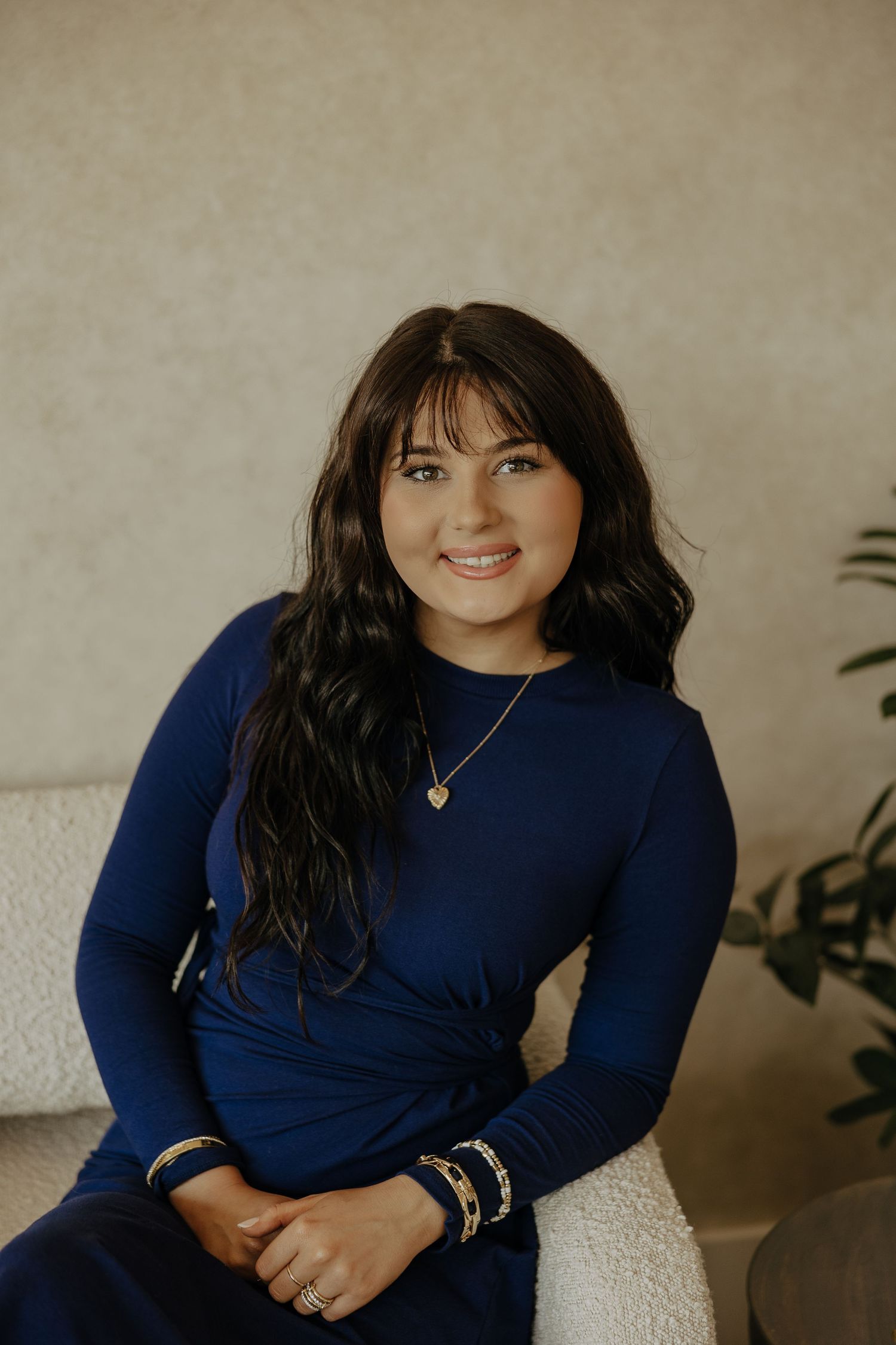 Professional portrait in a navy blue sweater with long dark hair and jewelry against a neutral background.