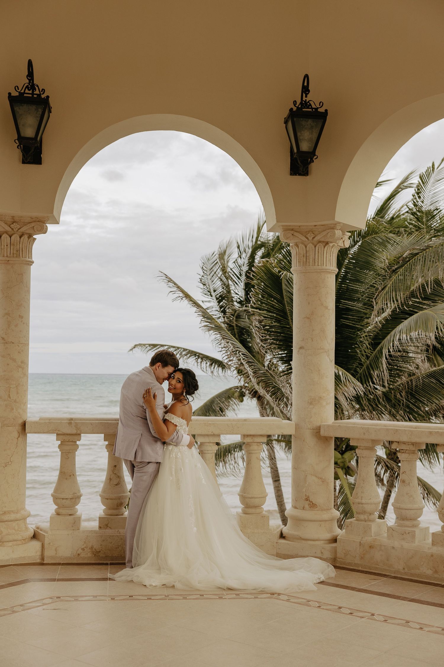 A romantic wedding couple embraces on an elegant white stone balcony overlooking a tropical beach at sunset.