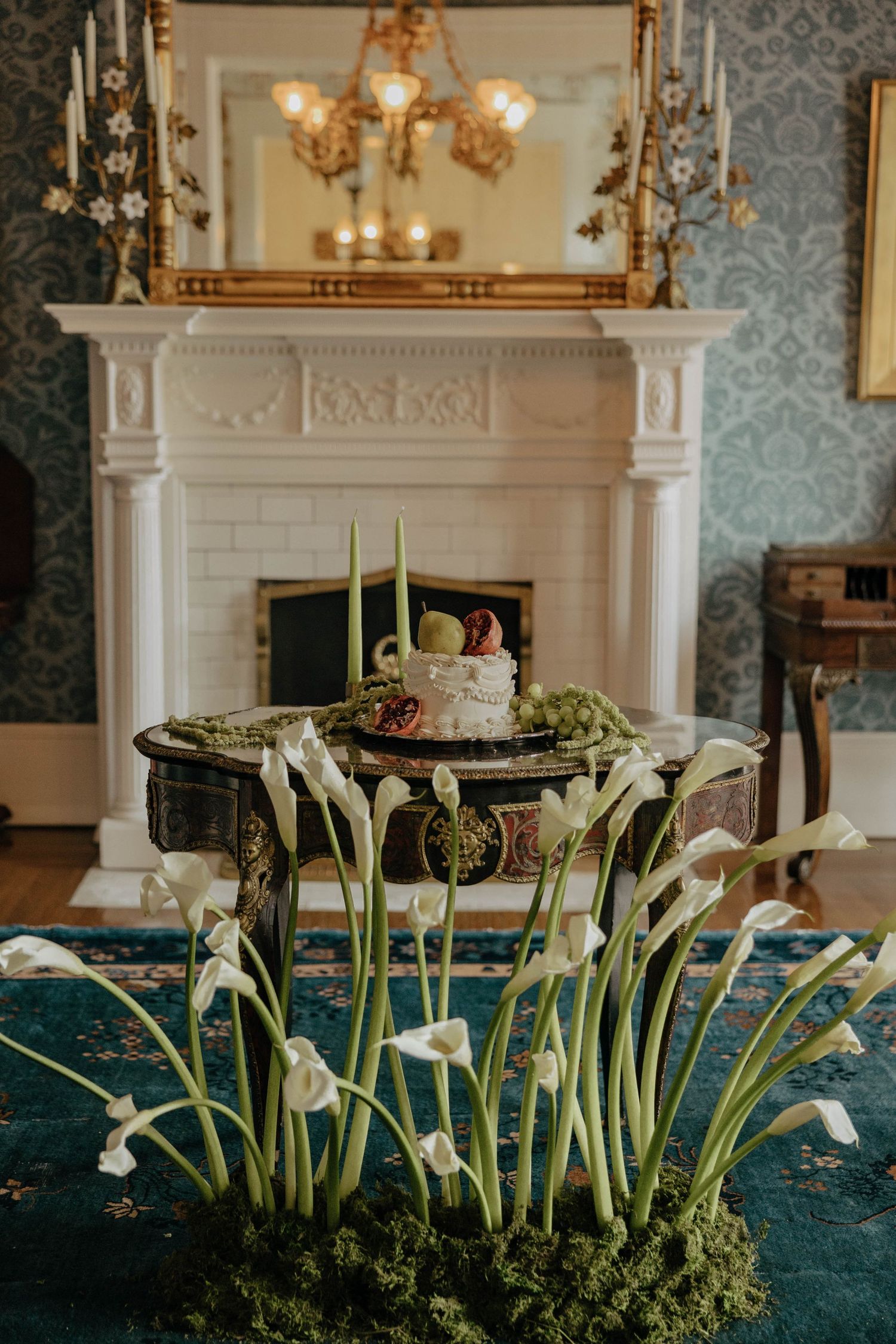 Elegant wedding cake display with white calla lilies on teal tablecloth in front of ornate white fireplace and mirror.
