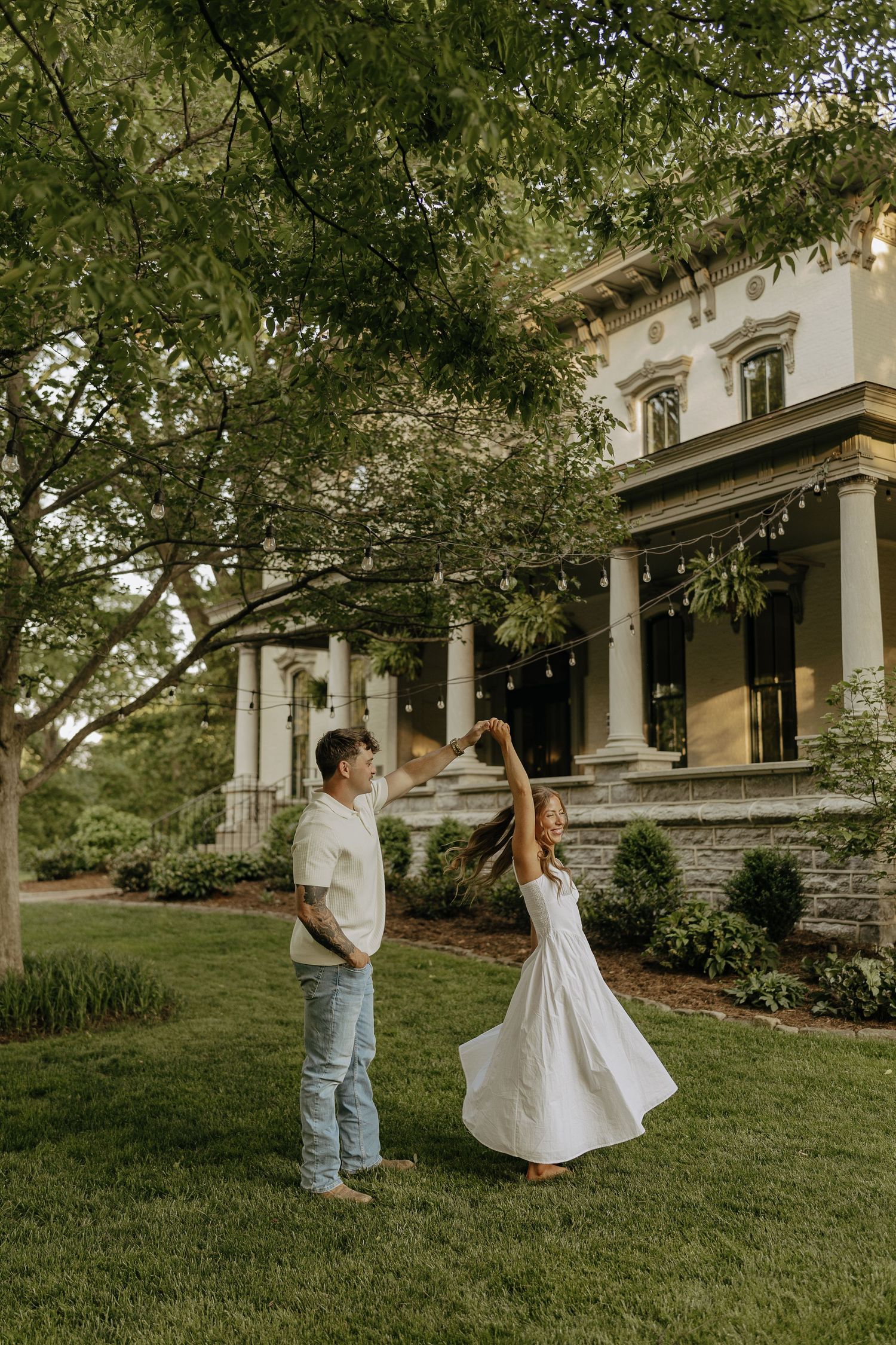 Couple dancing on green lawn in front of an elegant historic mansion with columns and string lights.