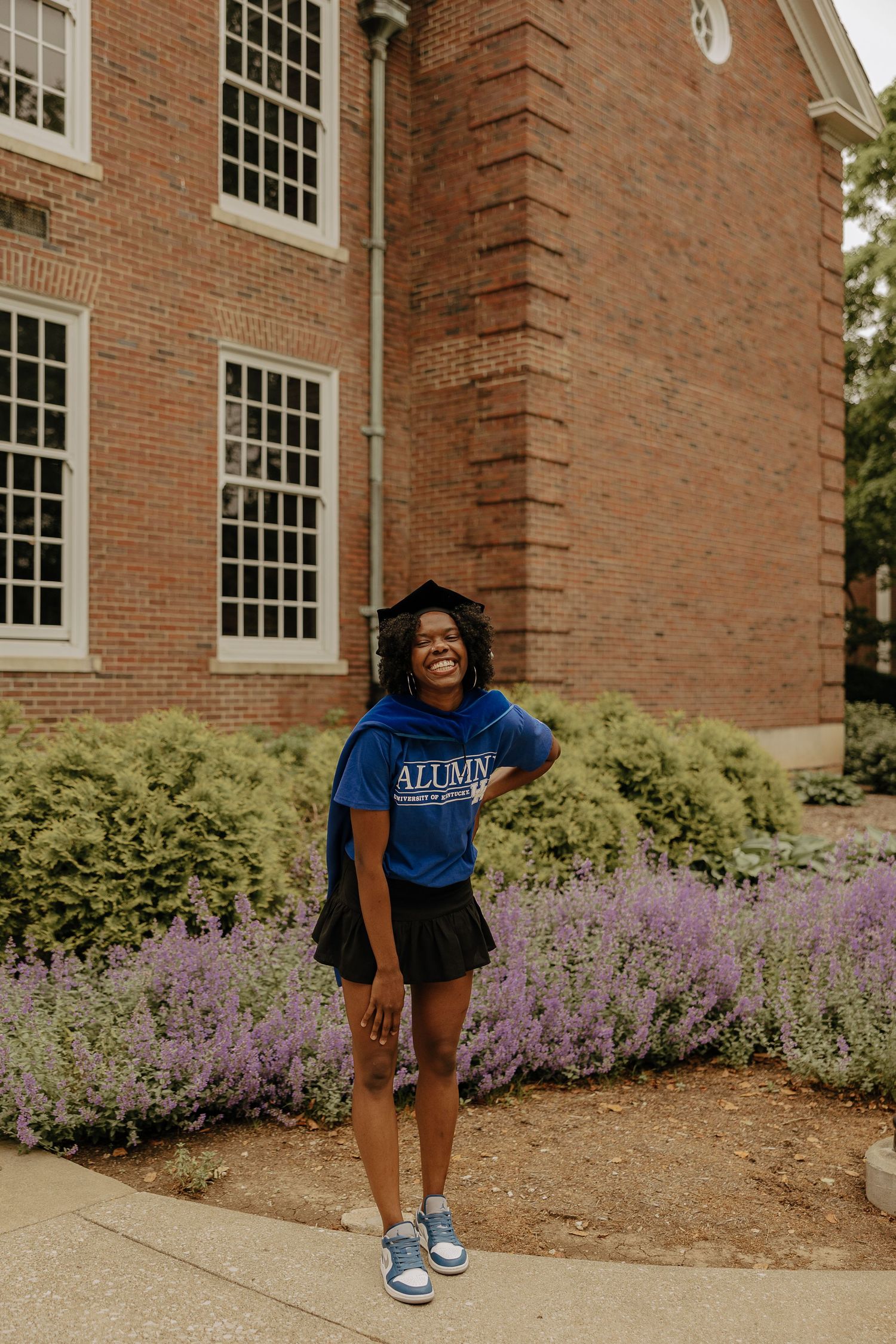 Blue alumni sweatshirt and black shorts ensemble photographed near purple flowers and brick building.