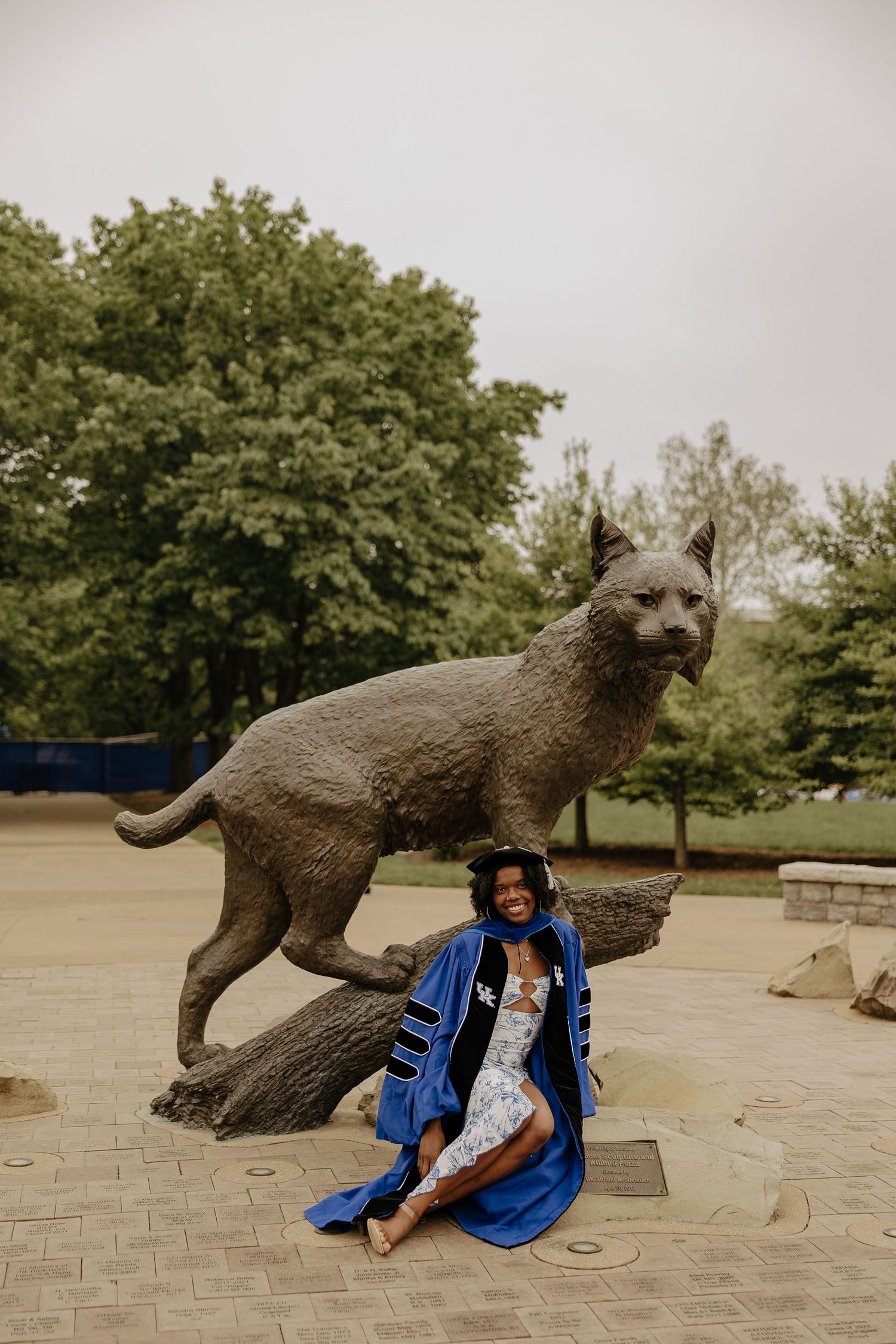 Graduate poses playfully with large wildcat statue on campus grounds wearing academic regalia.