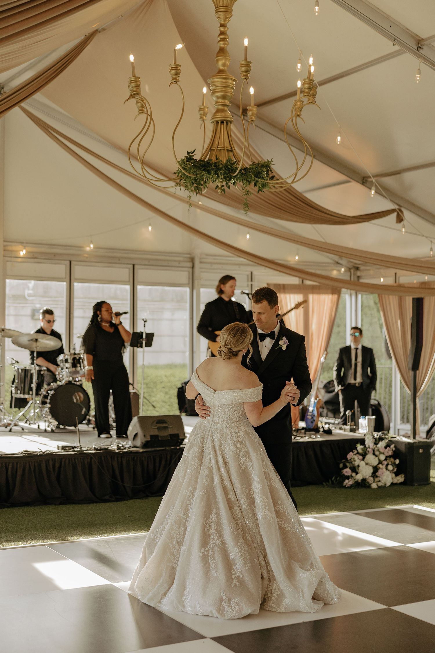 A wedding reception in a white tent with elegant chandeliers and draped fabric decor.