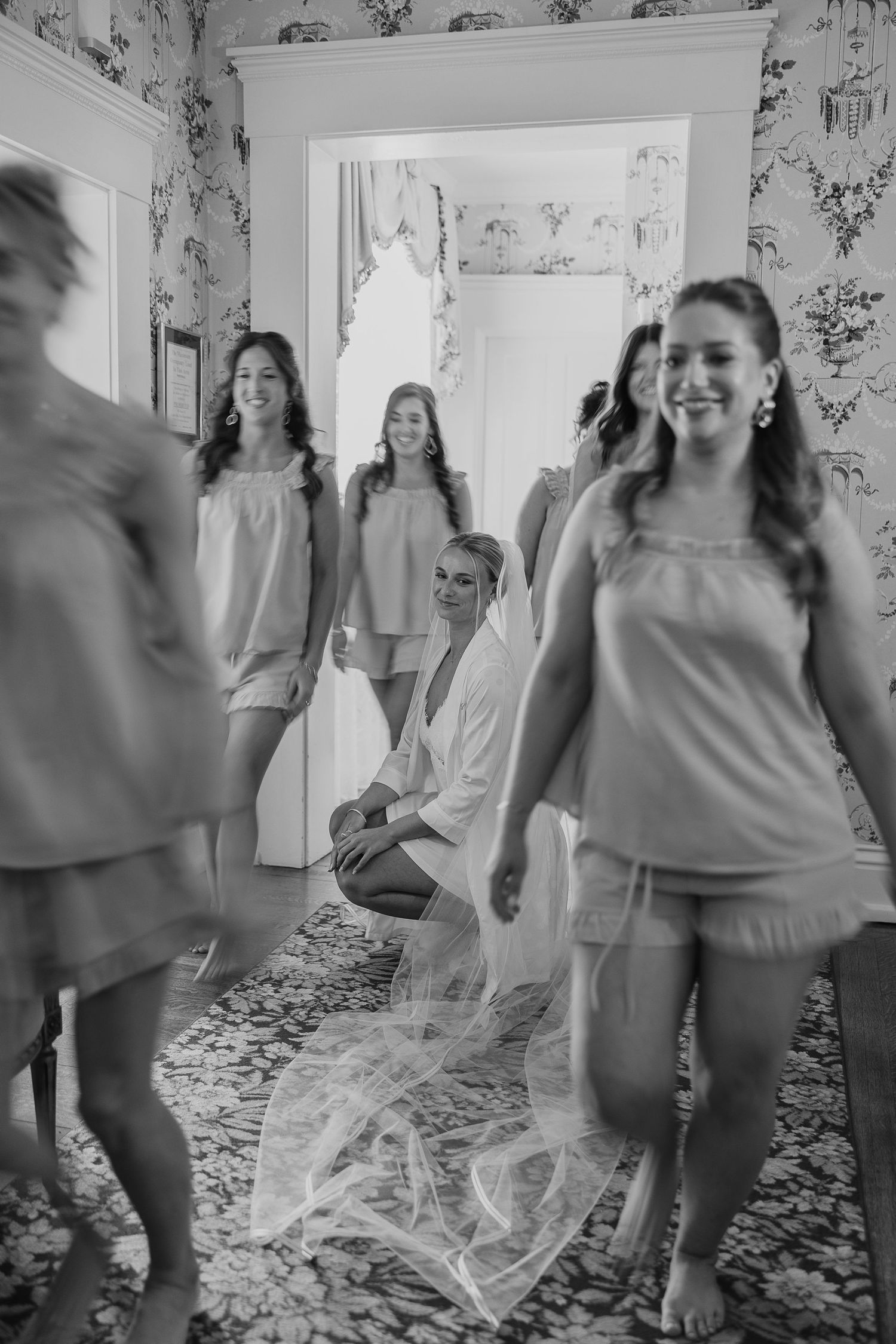 Bride kneeling with veil while bridesmaids in matching pajamas celebrate in an elegant wallpapered room.